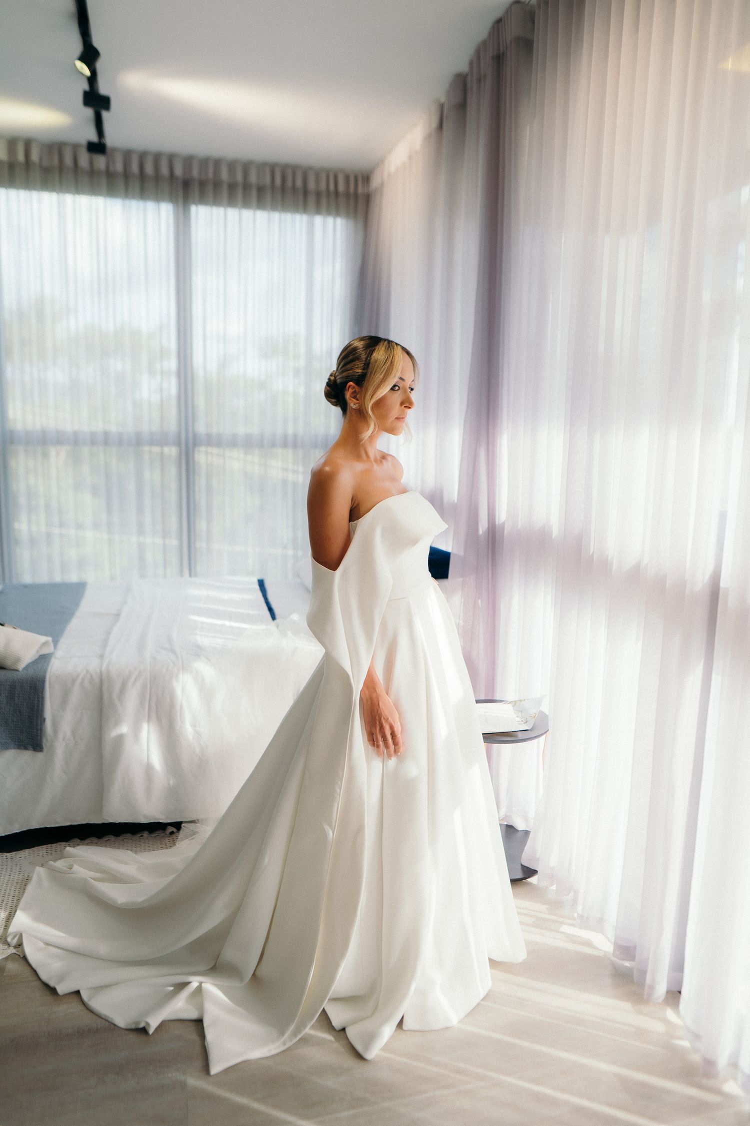 Bride in strapless white ball gown standing by large window in bright hotel room.