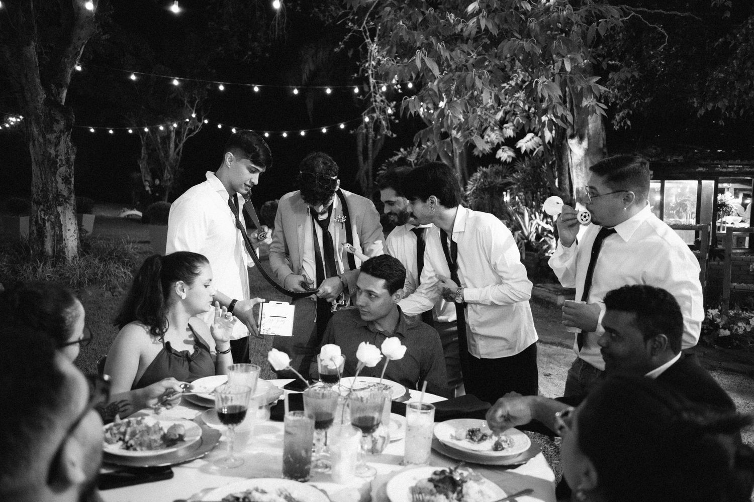 Wedding guests gathered around table under string lights during outdoor evening reception.
