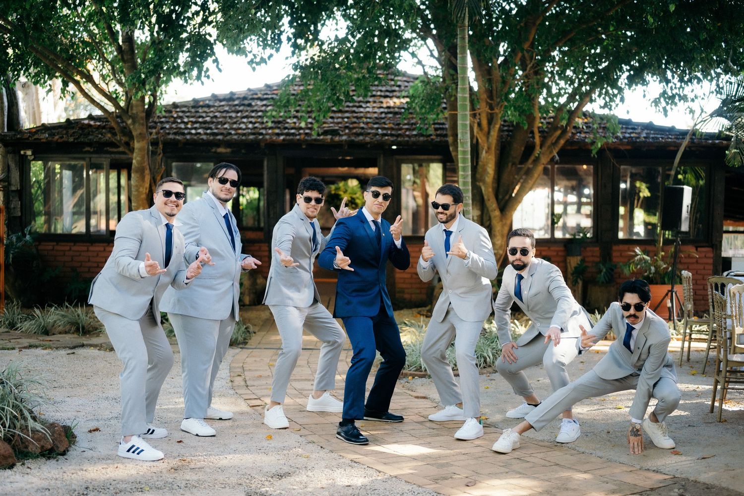 Groom in blue suit poses with groomsmen in gray suits and sunglasses, striking fun poses outdoors at a wedding venue.