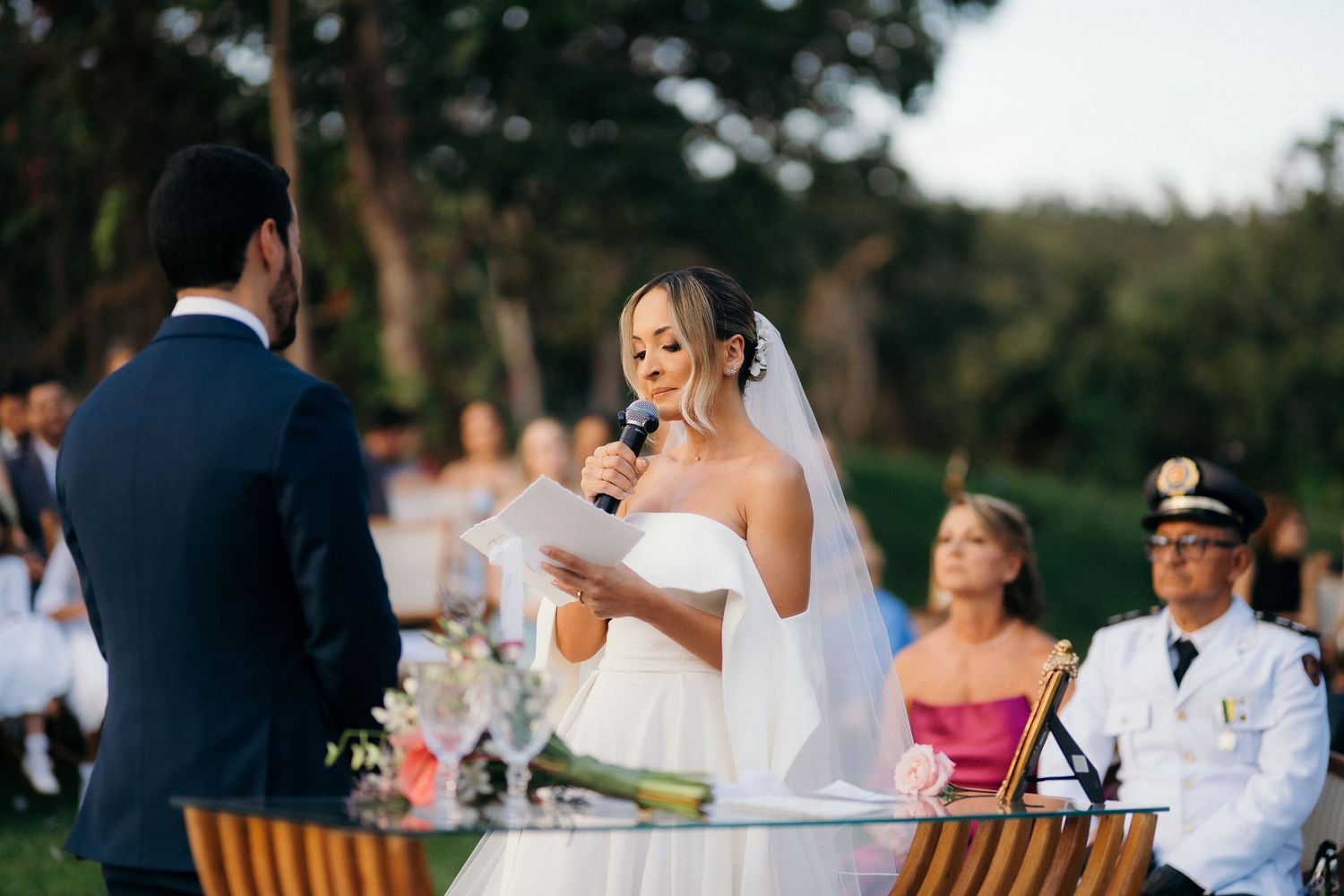 Bride in white gown reading vows into microphone at outdoor wedding ceremony.