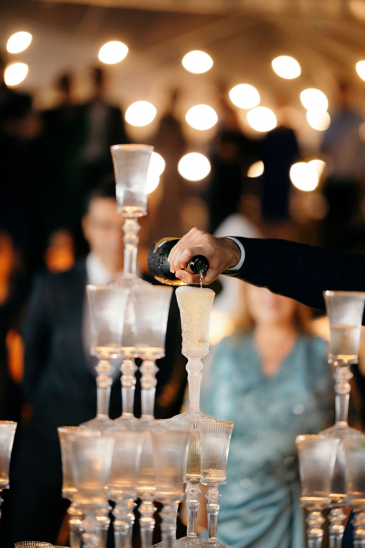 A waiter pours champagne into crystal glasses stacked in a pyramid tower at an elegant event celebration.