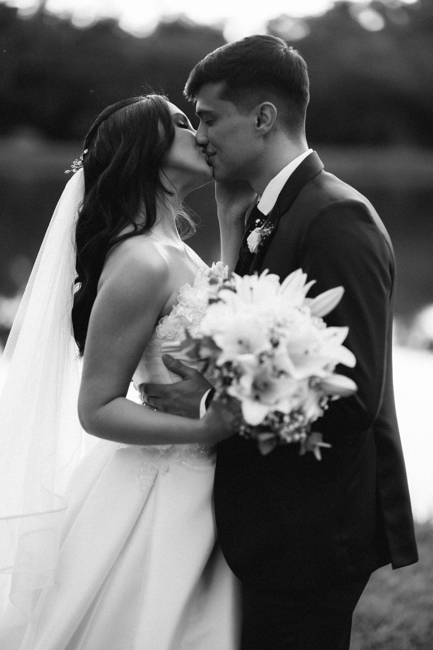 Black and white photo of bride and groom sharing a kiss holding white lily bouquet.
