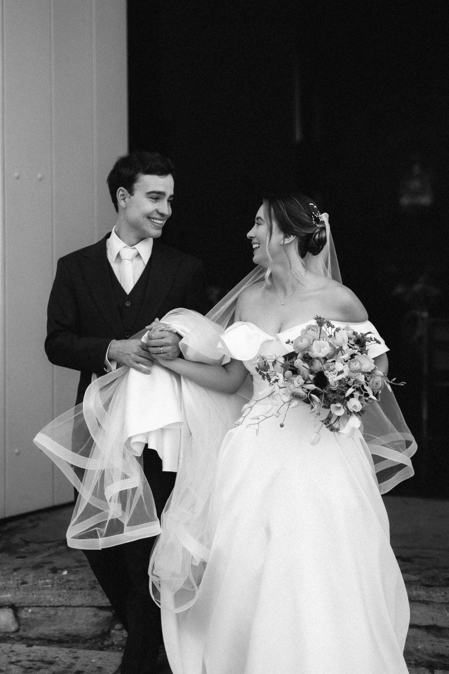 Black and white photo of a smiling bride and groom holding hands and walking together after their wedding ceremony.