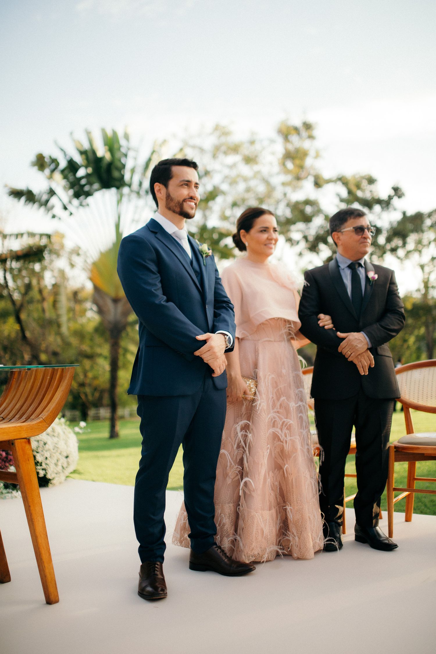 Bride in pink feathered gown walks down aisle with groom in blue suit at outdoor wedding ceremony.