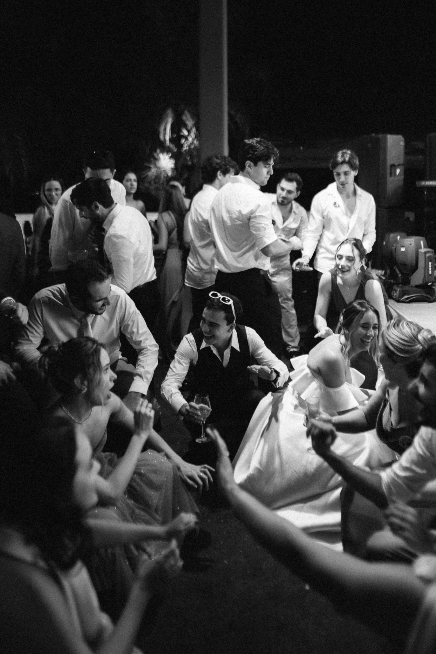 Black and white photo of wedding guests sitting on the floor celebrating with the bride at a lively reception.