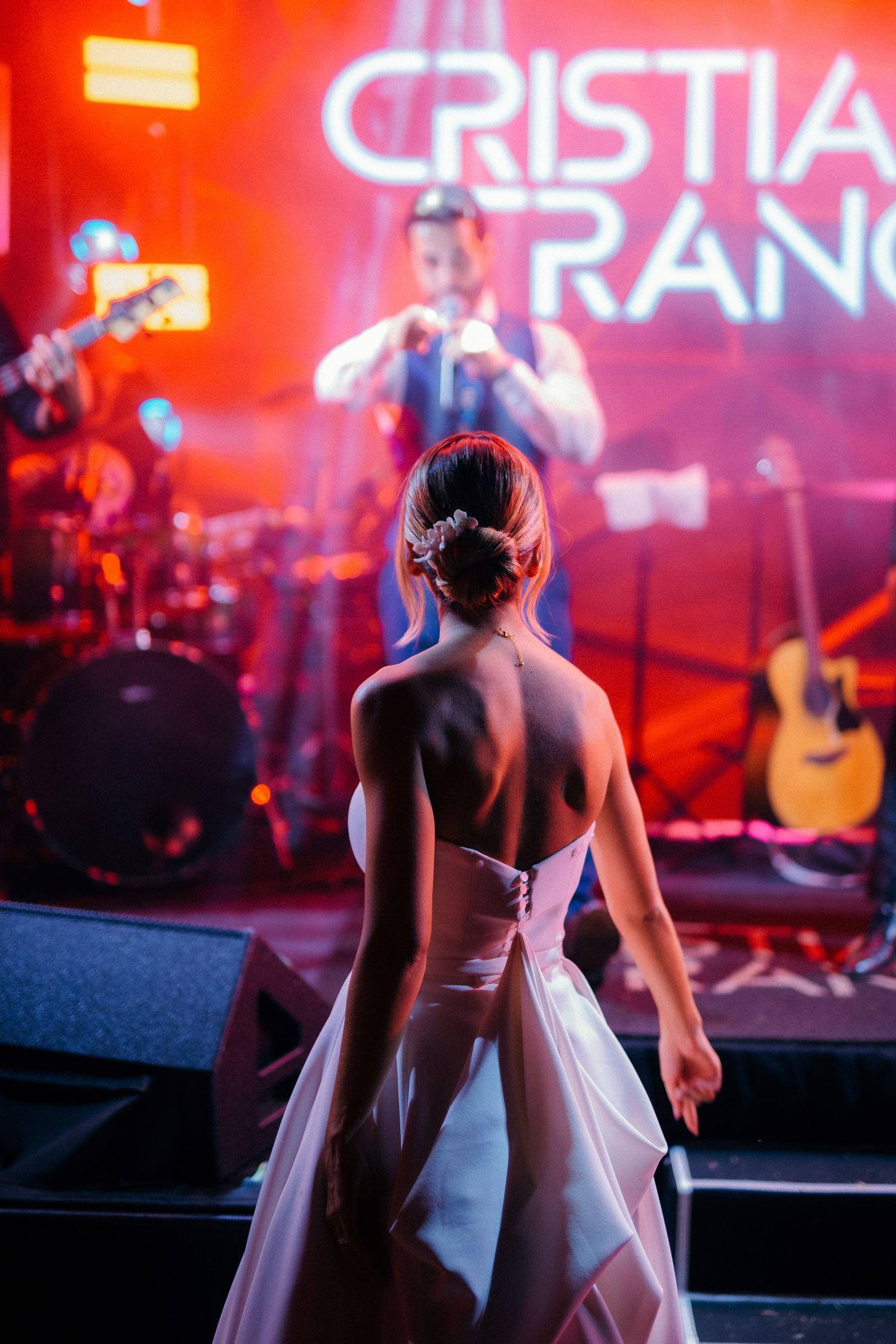 Bride in backless white gown watches live band perform on stage at wedding reception.