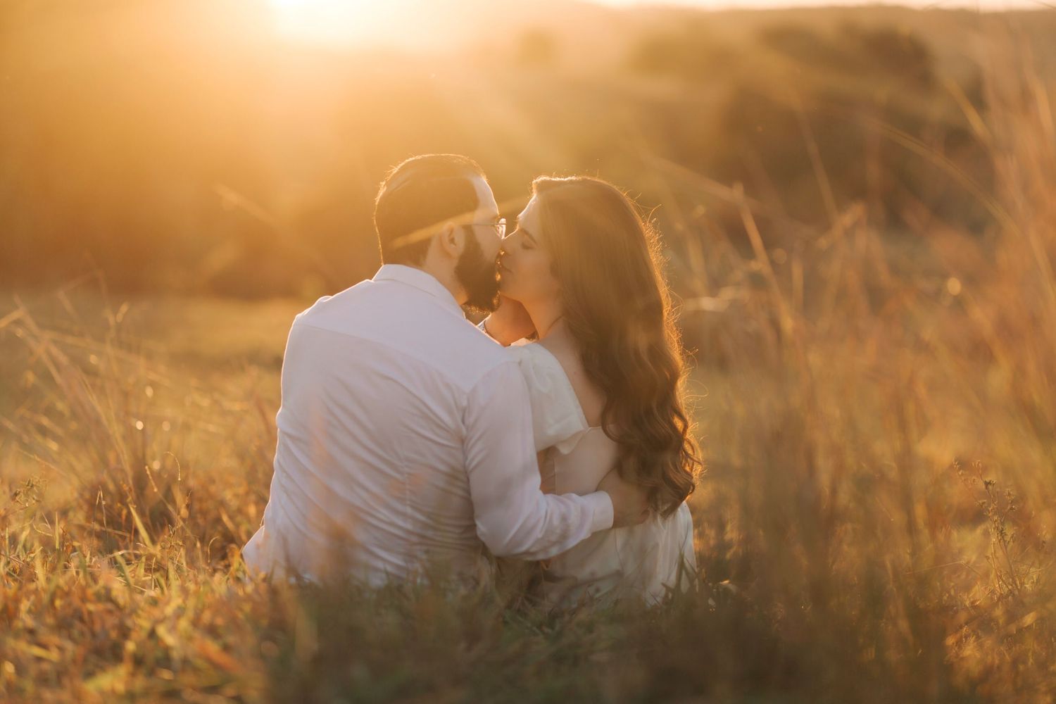 A couple shares a romantic kiss while sitting in a golden sunlit field during a warm sunset.