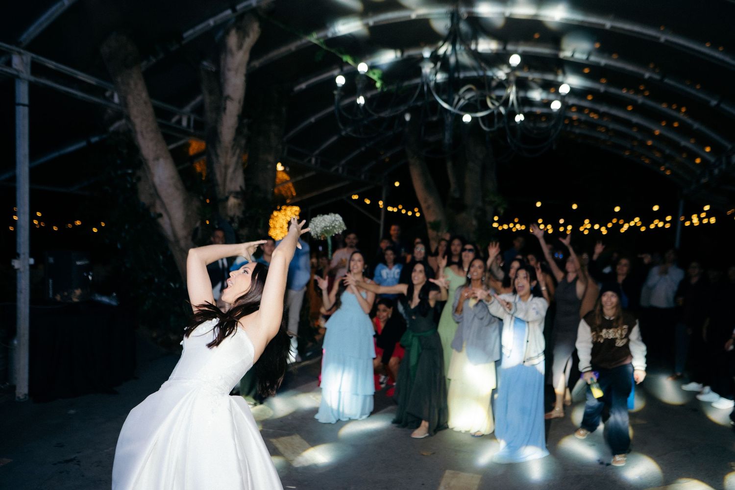 Bride tossing bouquet to excited guests under string lights at outdoor wedding reception.