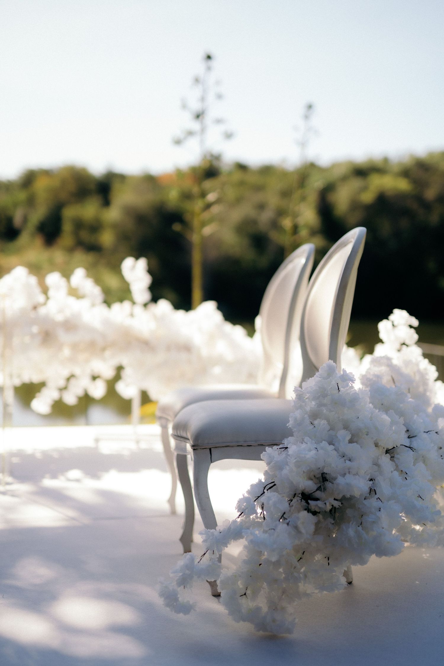 Elegant white wedding chairs and floral bouquet on white stage at outdoor ceremony venue.