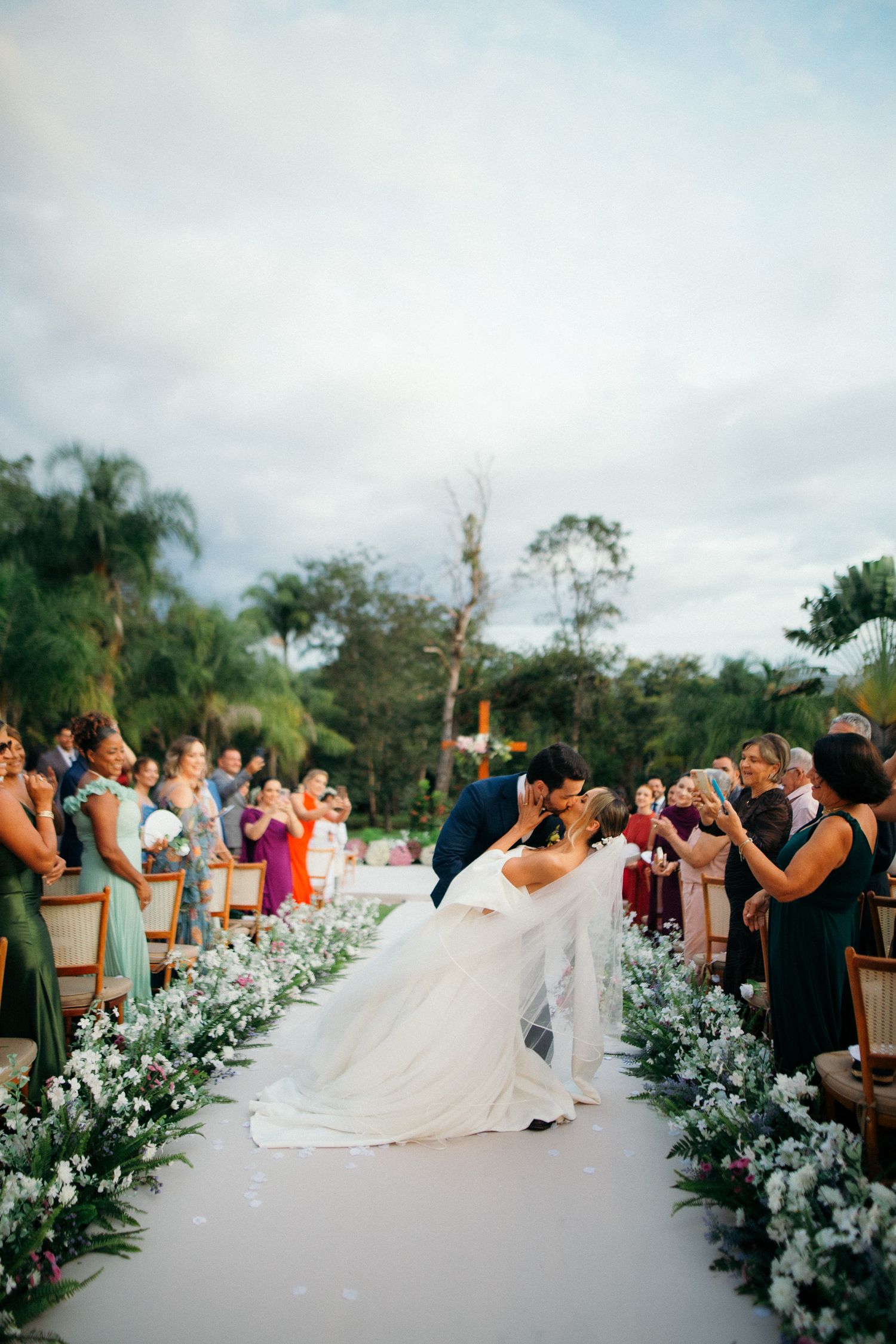 Bride and groom share romantic dip kiss in wedding aisle surrounded by floral arrangements and guests.