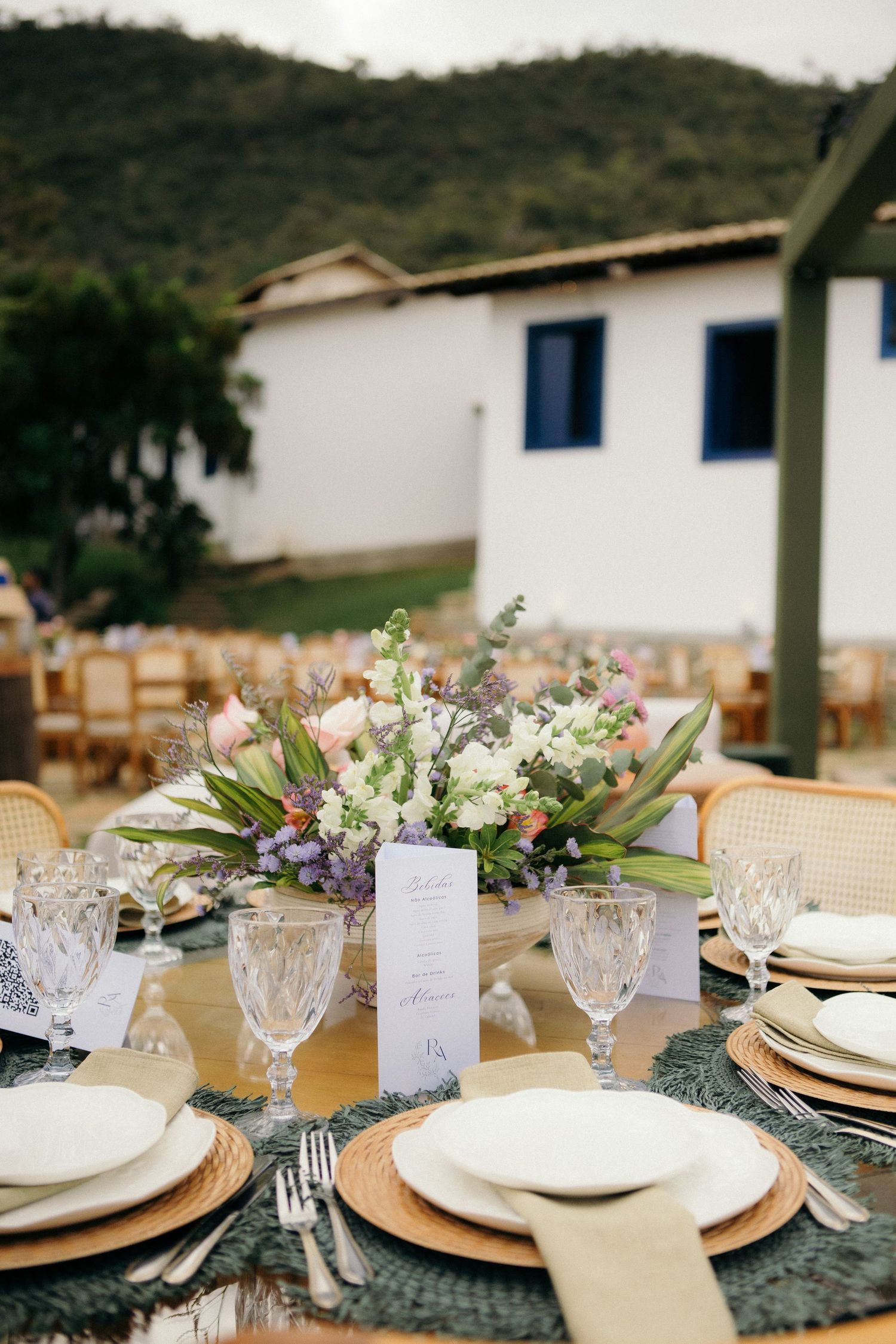 Elegant outdoor wedding reception table set with crystal glasses, floral centerpiece, and wicker chargers against a scenic backdrop.
