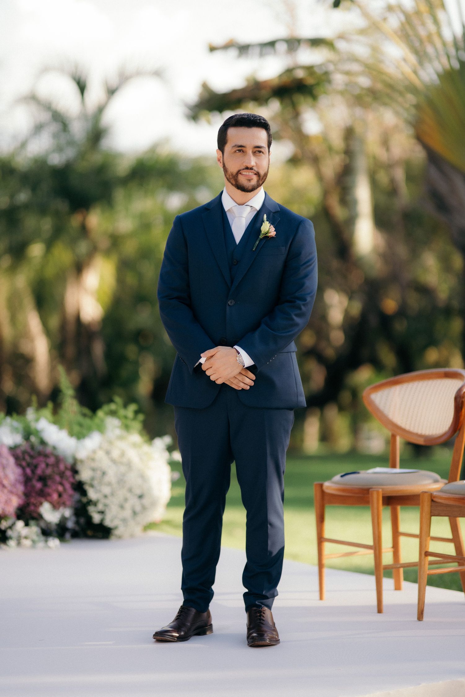 Groom in navy blue suit with boutonniere stands at outdoor wedding ceremony aisle.
