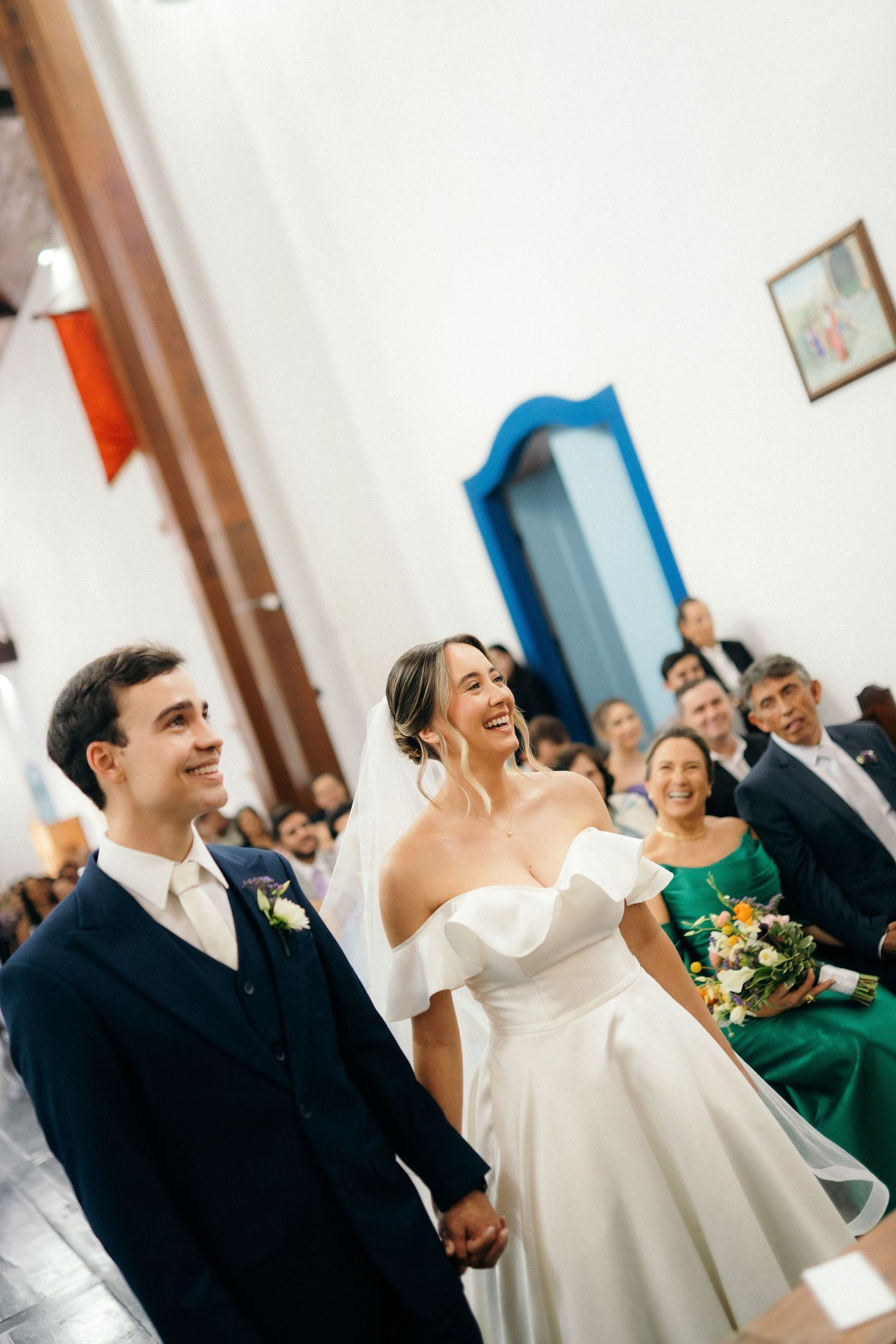 Joyful bride and groom holding hands and laughing during church wedding ceremony with guests.