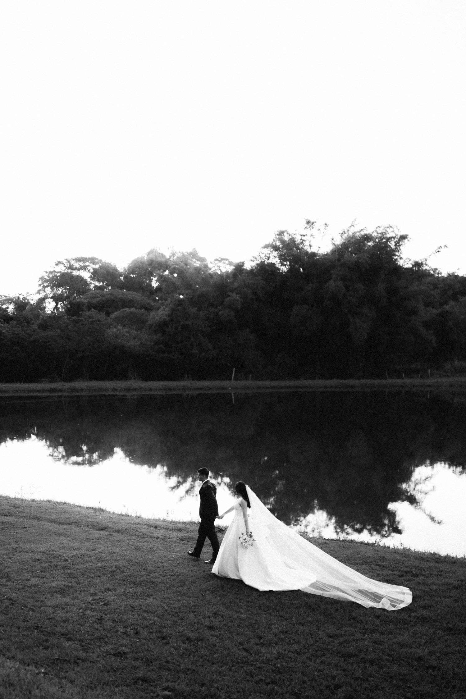 Black and white photo of a bride and groom walking hand in hand along a reflective lake at sunset.