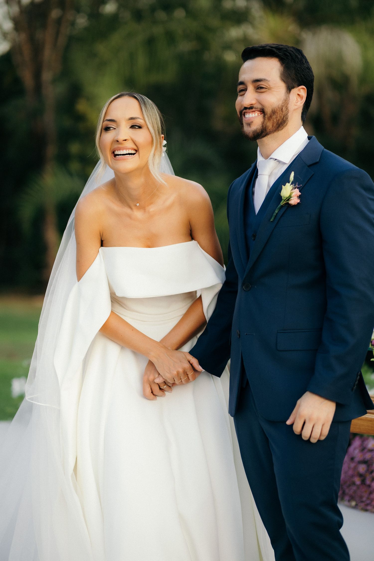 Joyful bride in off-shoulder white gown and groom in navy suit hold hands smiling during outdoor wedding ceremony.