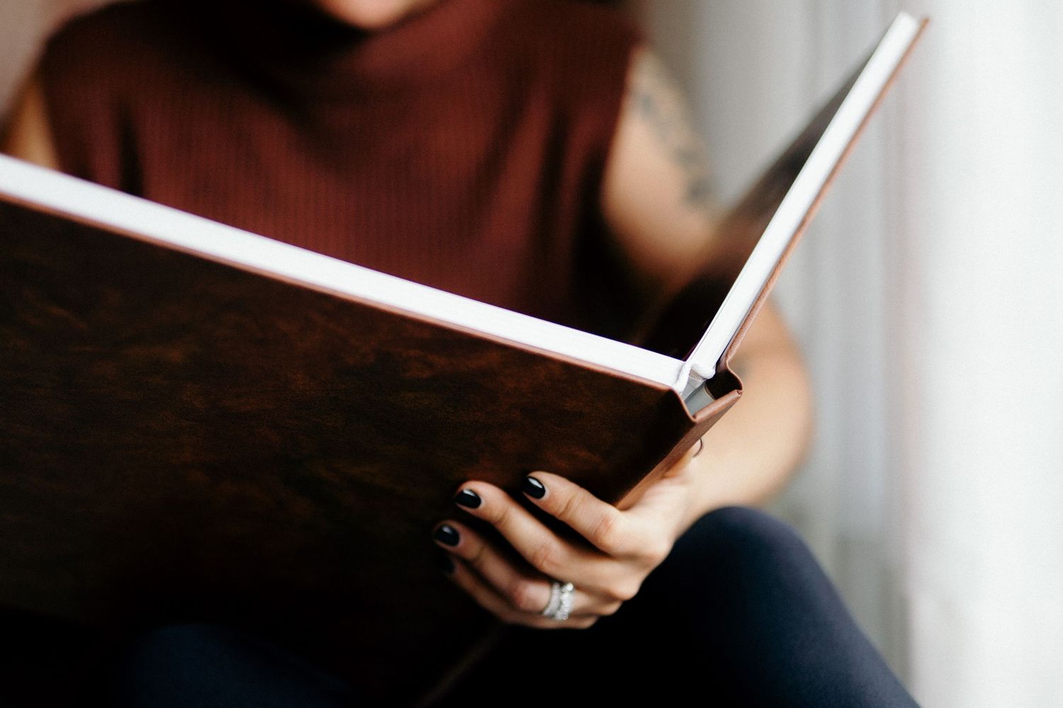 Woman with dark nails and ring holding open a large dark hardcover book or photo album indoors.