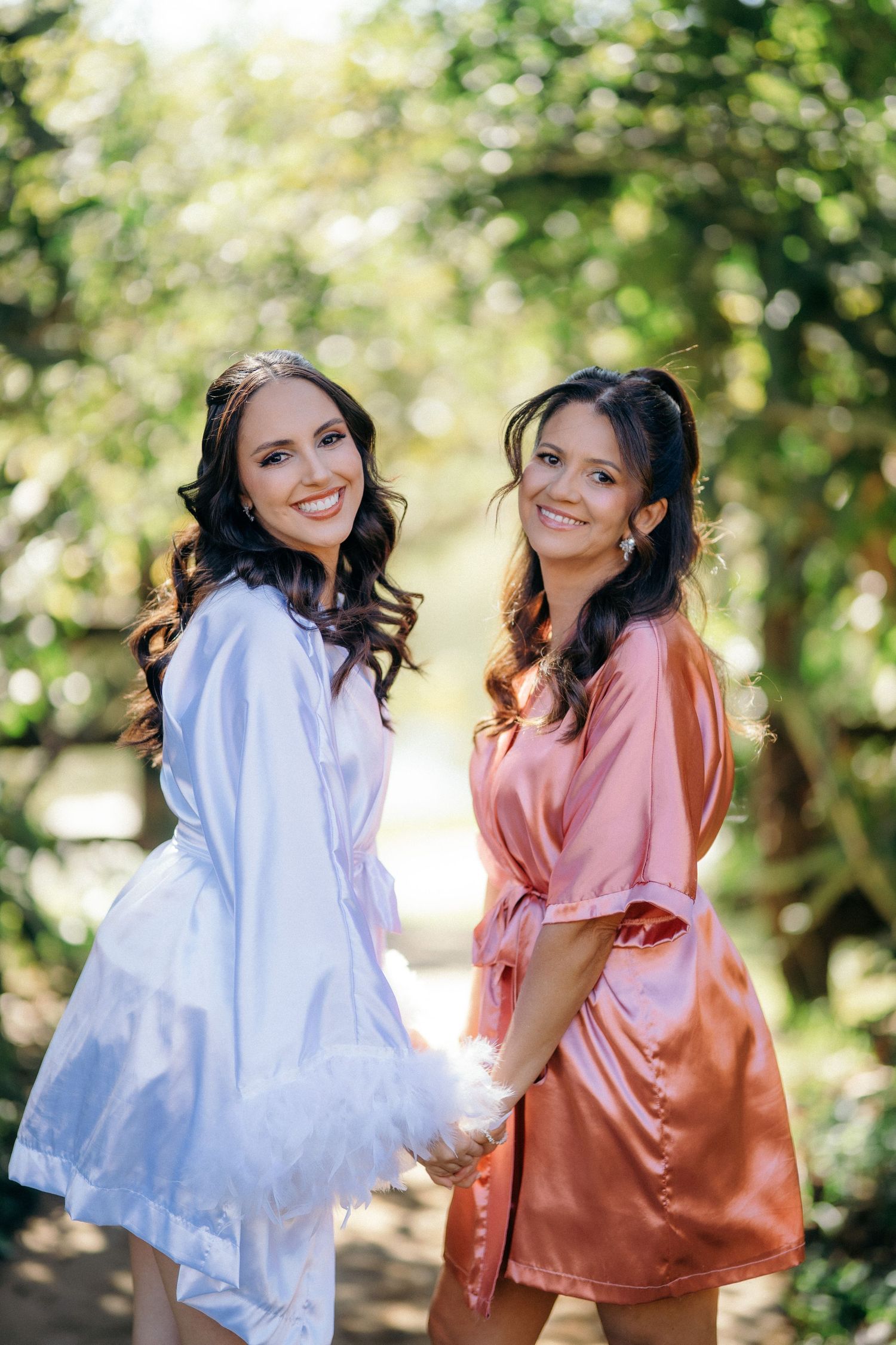 Two smiling women in satin robes holding hands in a sunlit garden setting.