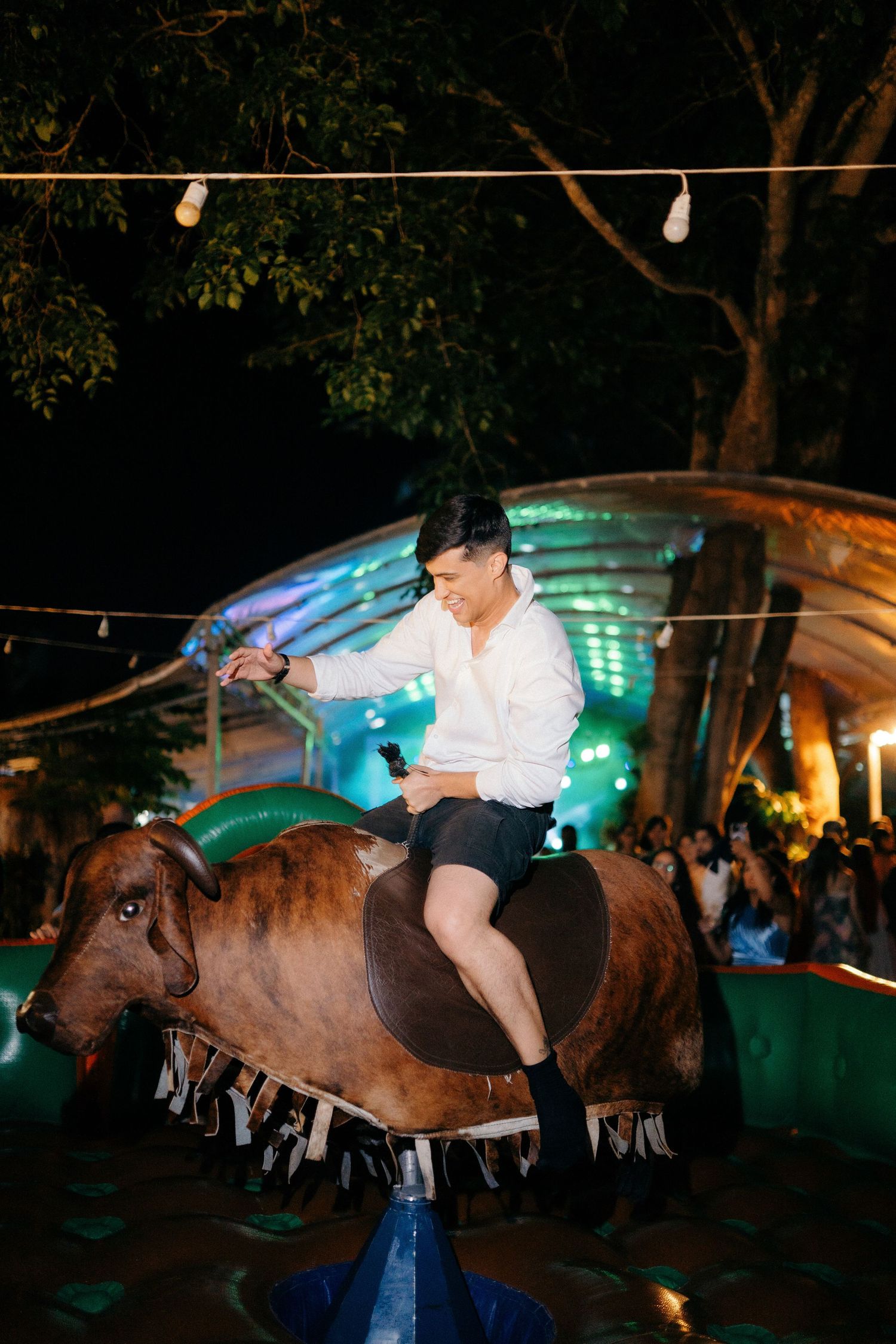Guest rides mechanical bull at lively outdoor wedding reception under string lights at night.