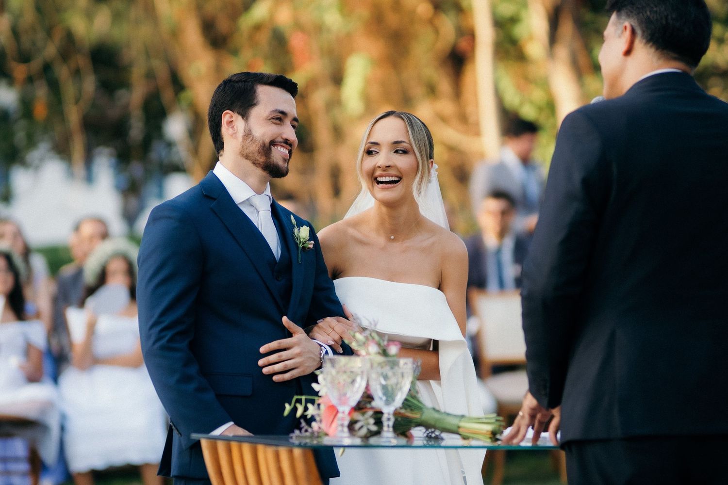 Joyful bride in off-shoulder gown and groom in navy suit laughing together during outdoor wedding ceremony.