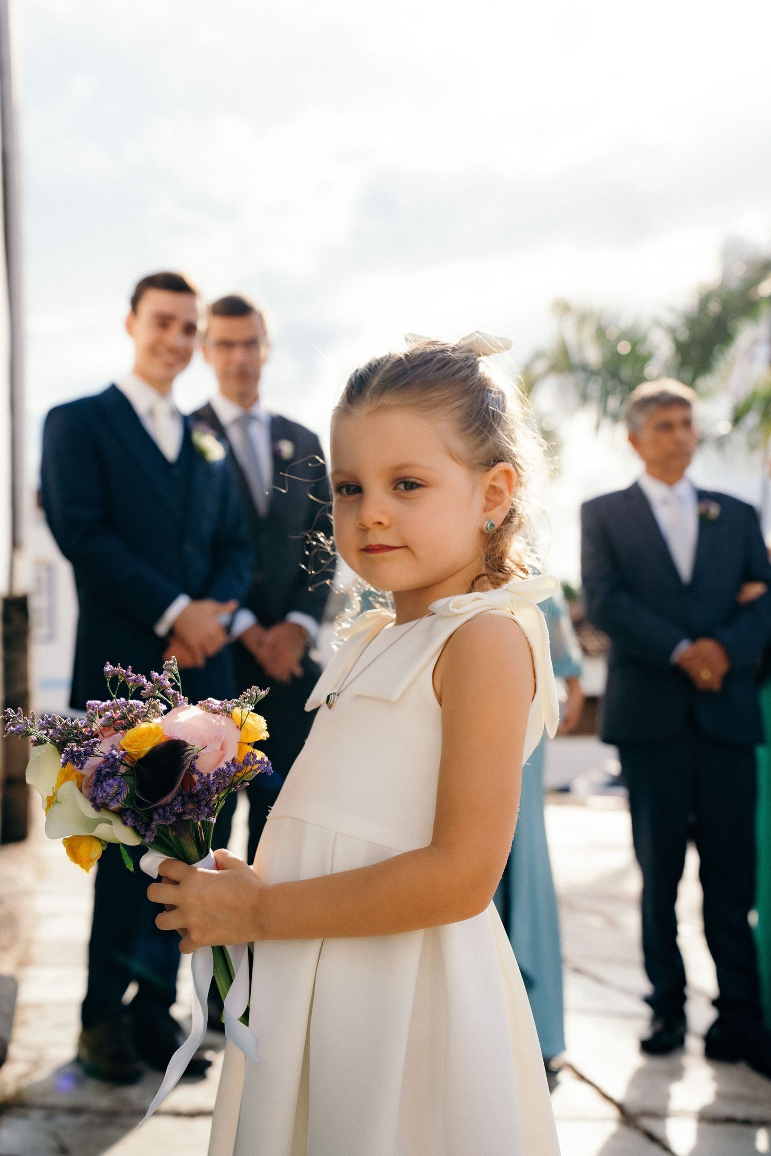 Young flower girl in white dress holding colorful bouquet with groomsmen standing behind her outdoors.