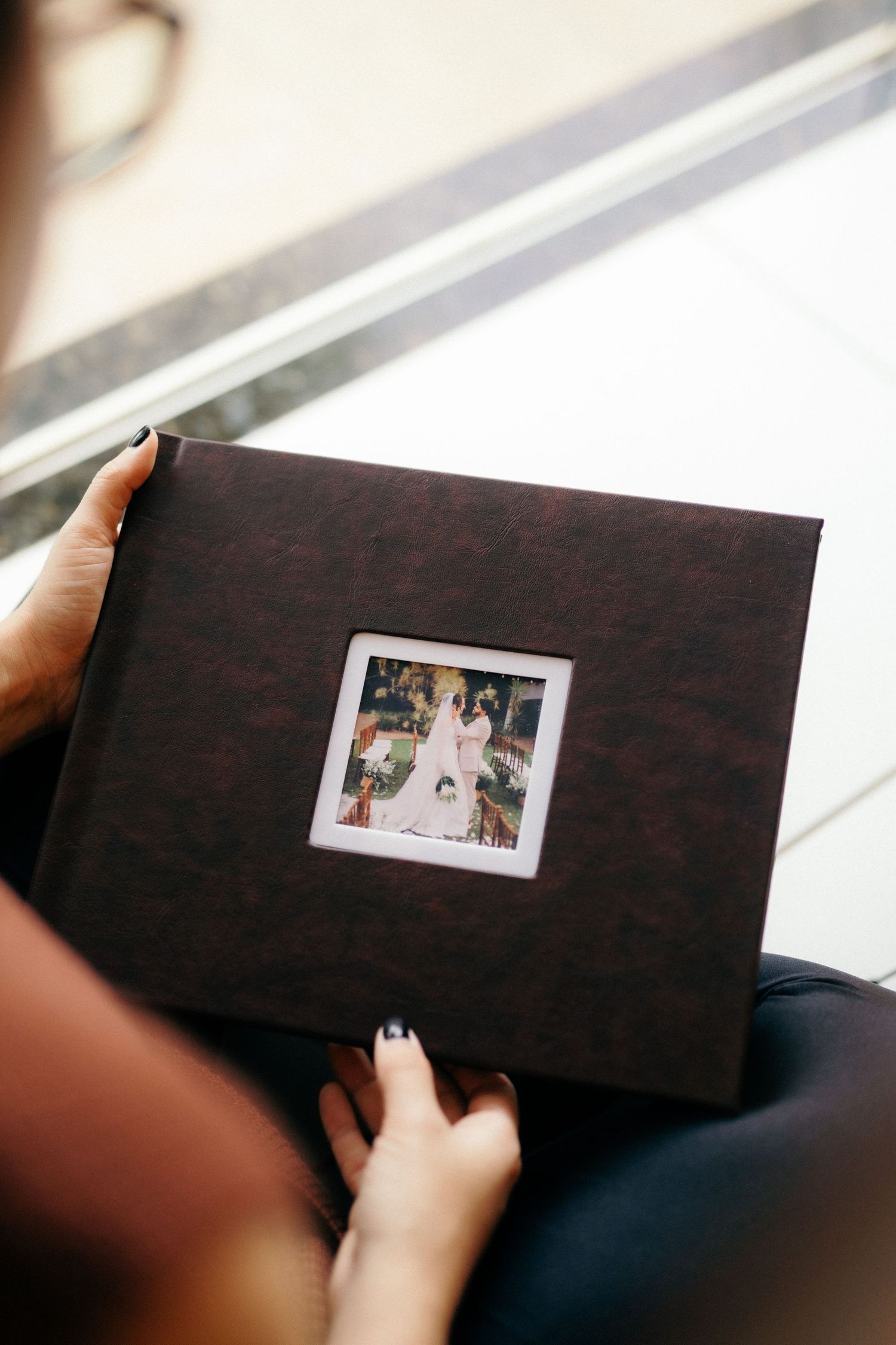 Person holding a dark brown leather wedding photo album with a small portrait window on the cover.