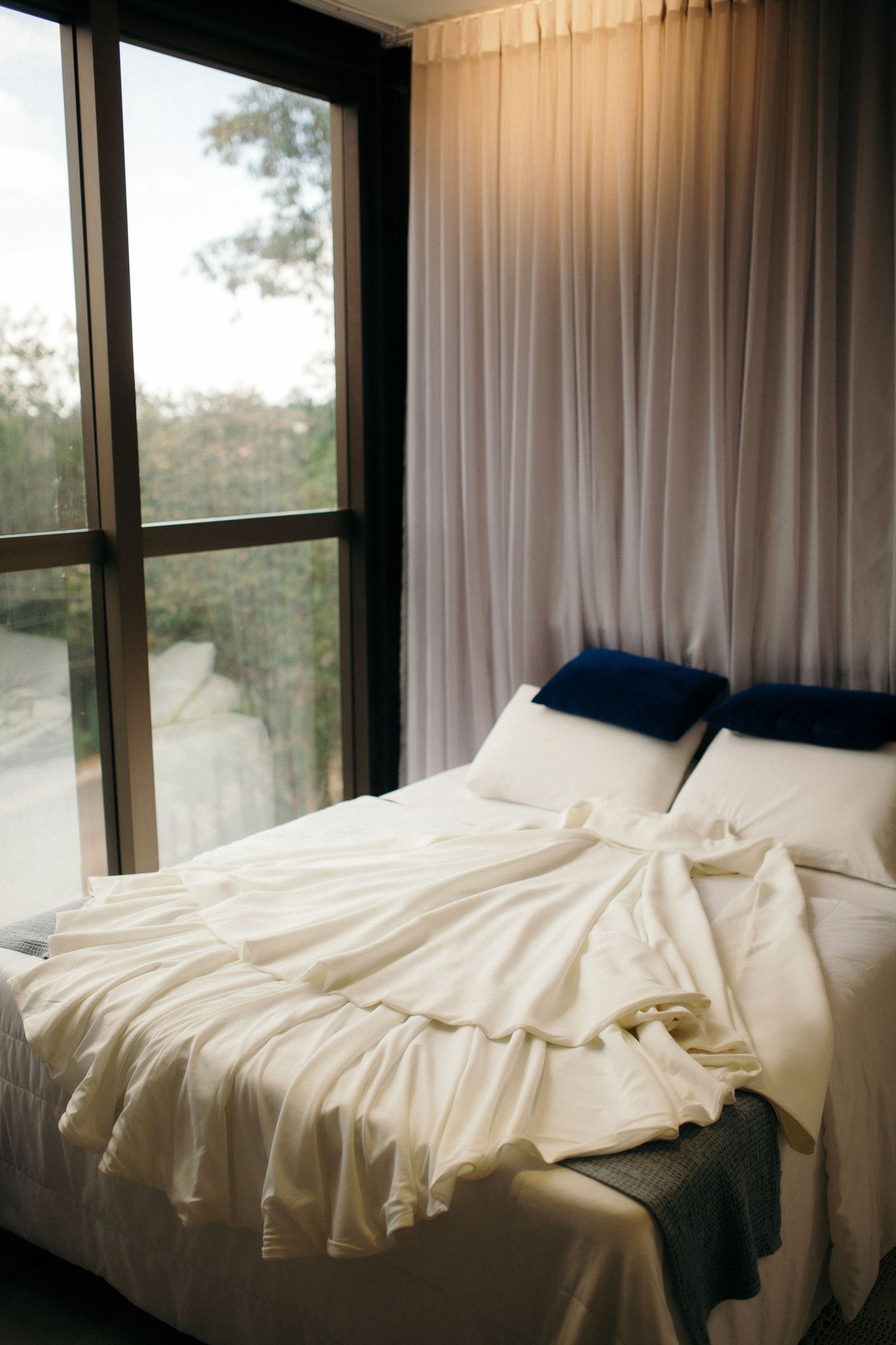 Sunlit bedroom with unmade white bedding, blue accent pillows, sheer curtains, and large windows overlooking lush greenery.