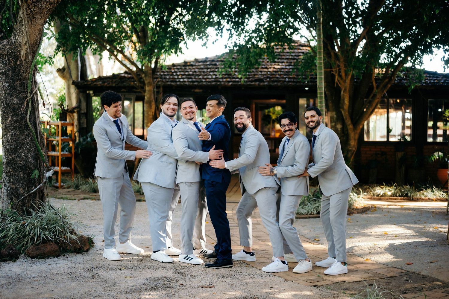 Groom in navy suit surrounded by groomsmen in light gray suits laughing together outdoors at a wedding venue.