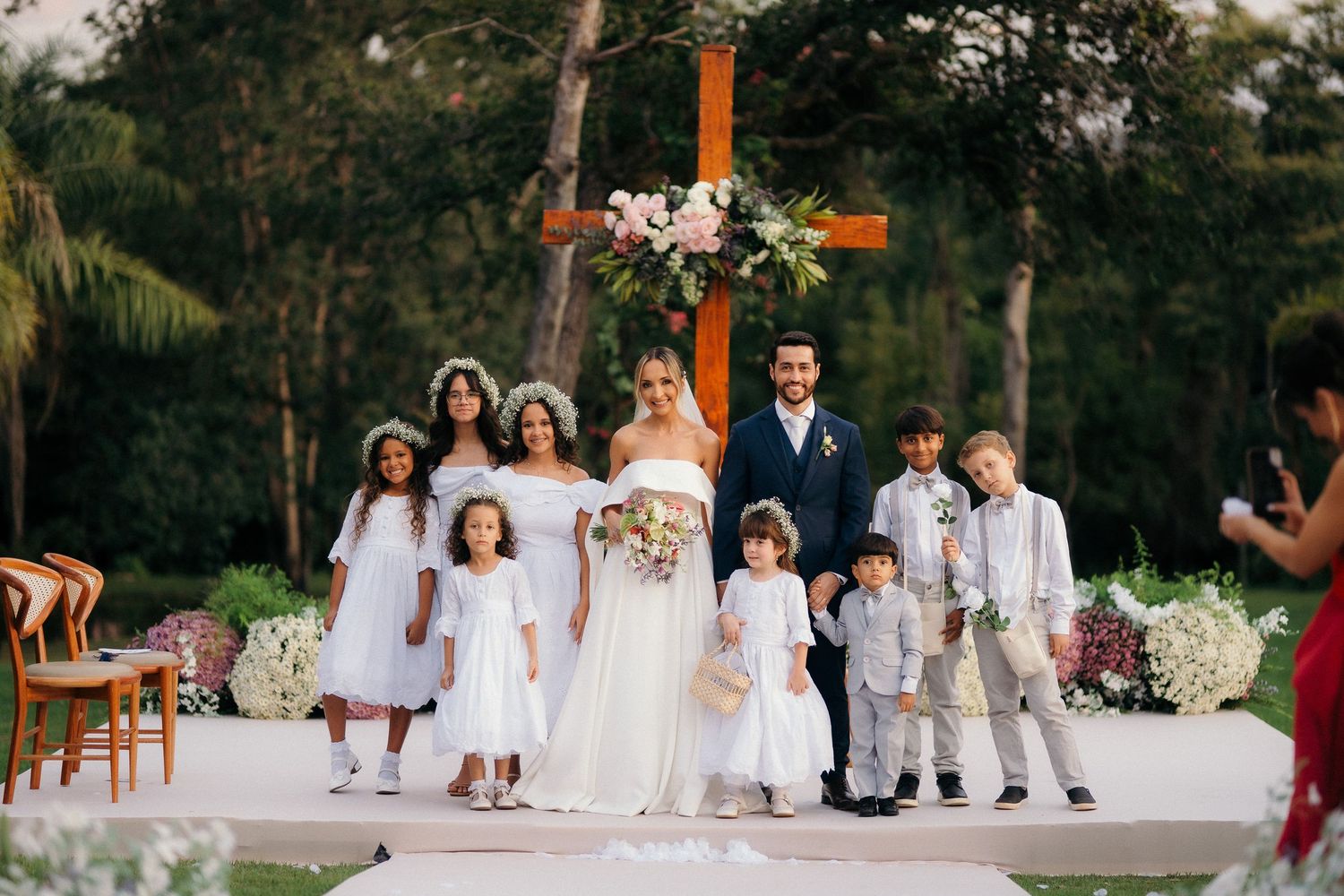 Bride and groom pose with flower girls and ring bearers at outdoor wedding ceremony with floral cross.