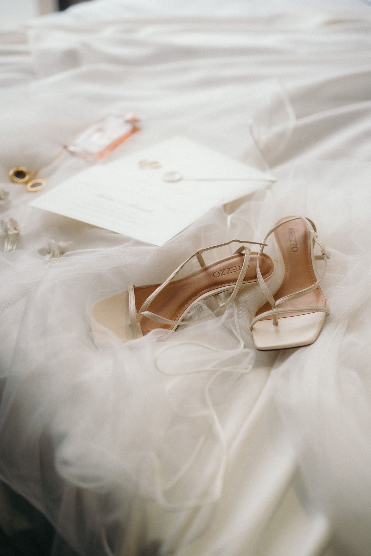 Bridal details flat lay with strappy heels, wedding invitation, rings, and perfume on white tulle.
