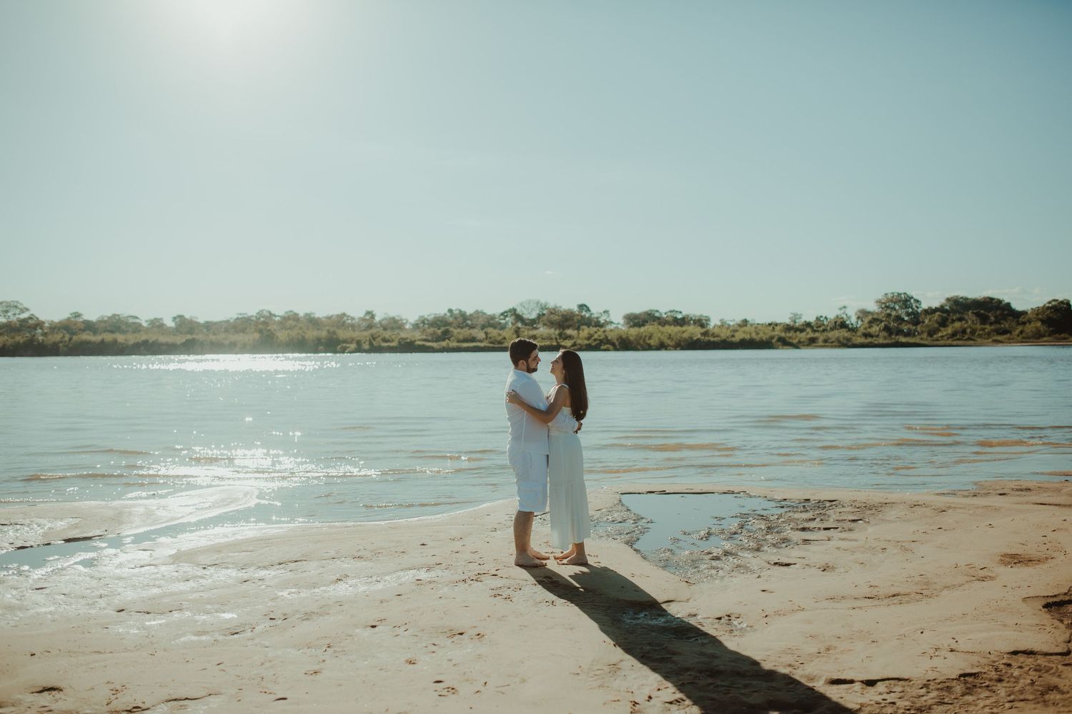 Happy couple in white outfits embracing on a sandy riverbank under bright sunny sky.