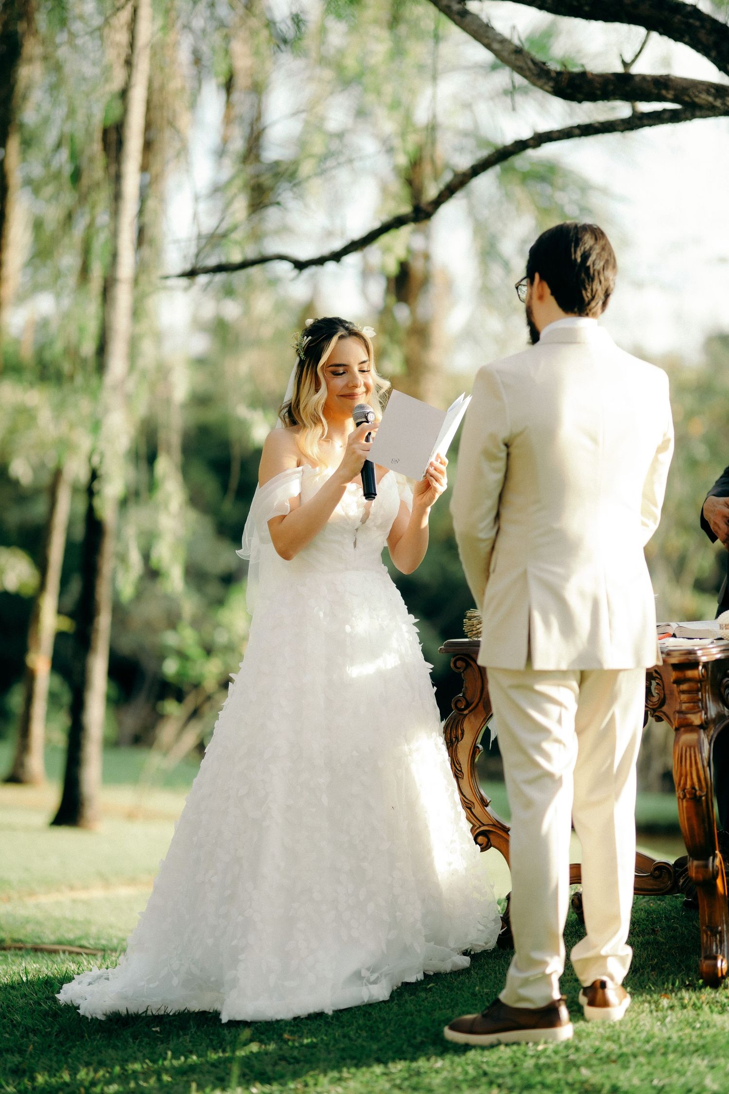 Bride reads wedding vows to groom in white gown at outdoor garden ceremony surrounded by lush green trees.