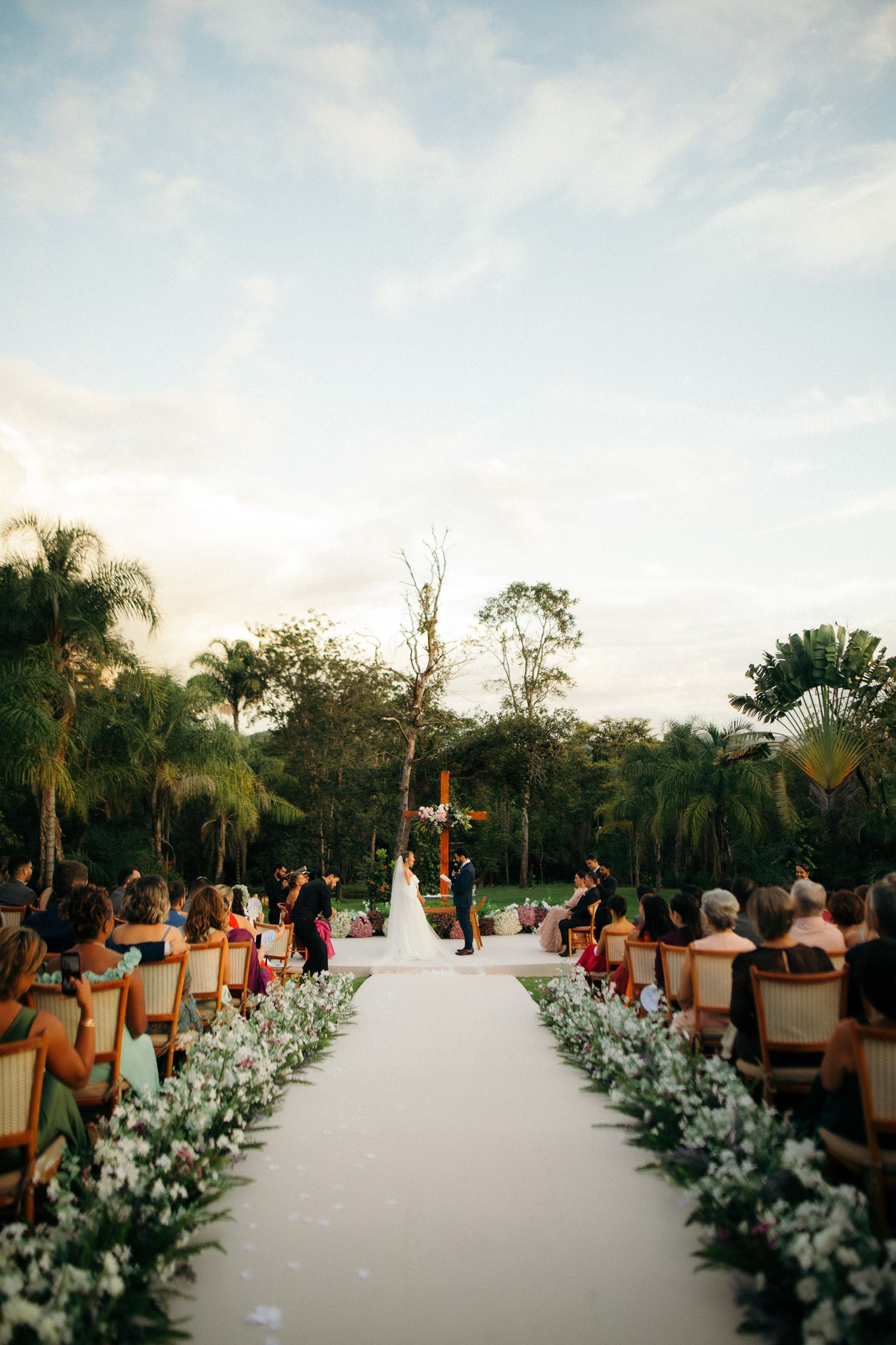 Outdoor wedding ceremony with white aisle flanked by flowers leading to couple at altar among palm trees.