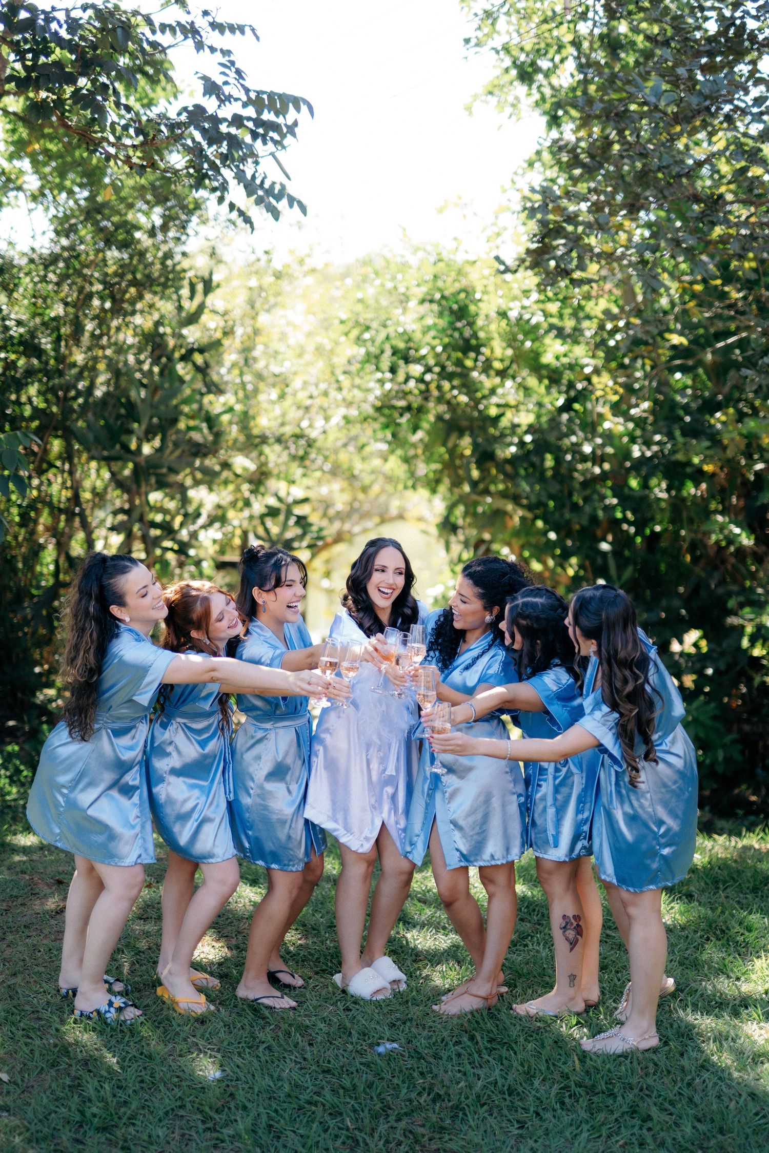 Bridesmaids in blue satin robes toasting champagne with bride in white robe outdoors in lush garden.