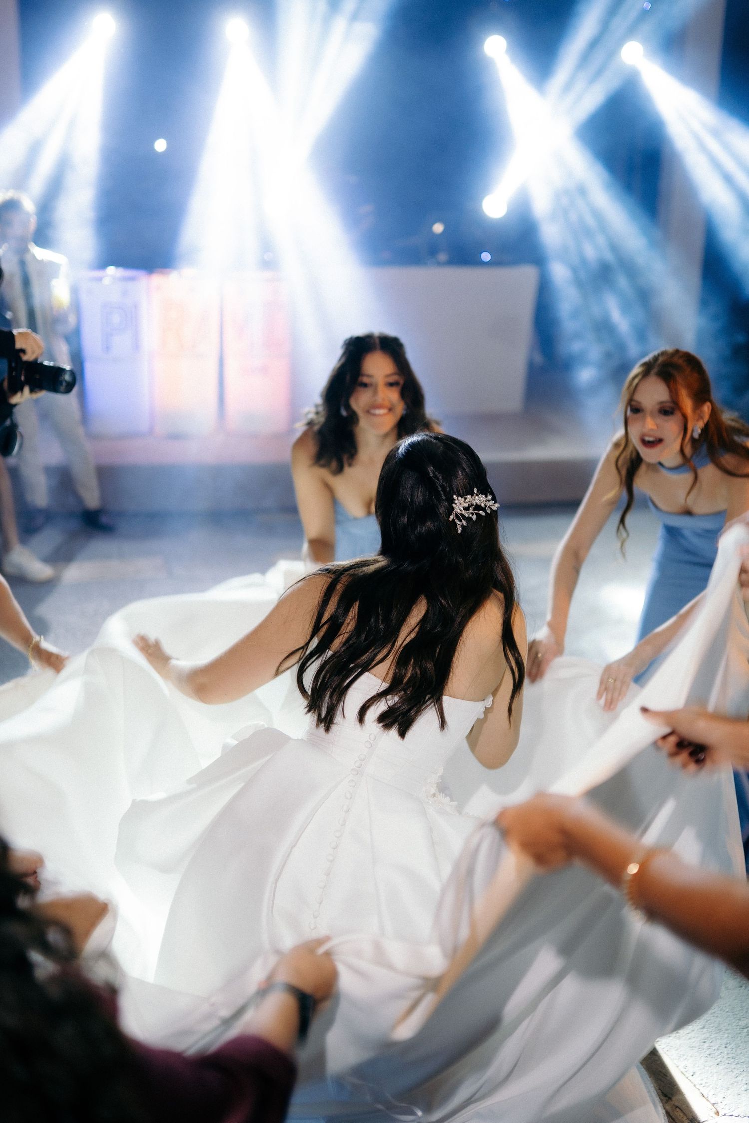 Bride in white gown dances joyfully with bridesmaids in blue dresses under dramatic stage lighting at wedding reception.