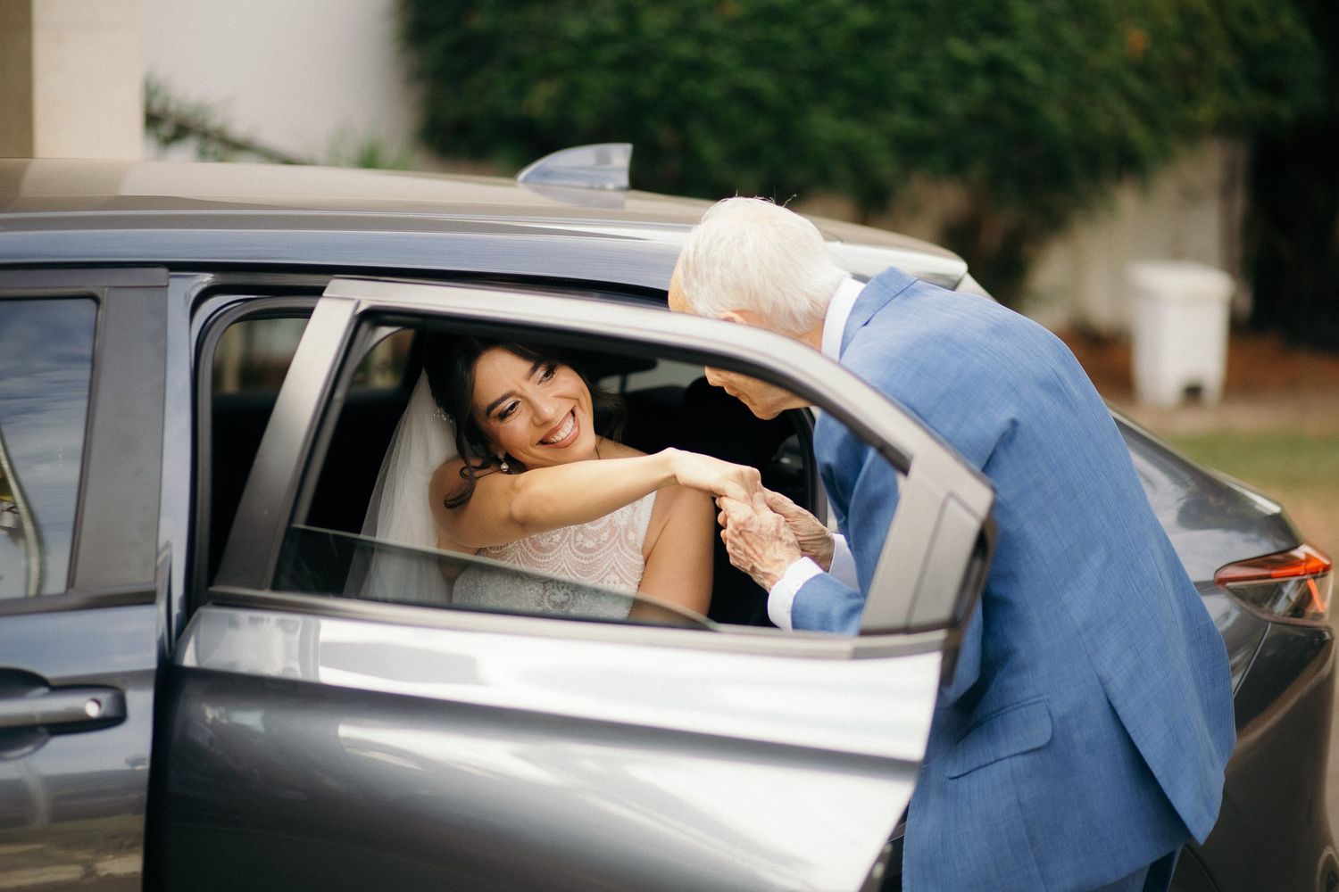 Elderly man in blue suit greeting smiling bride in white dress through car window at wedding.