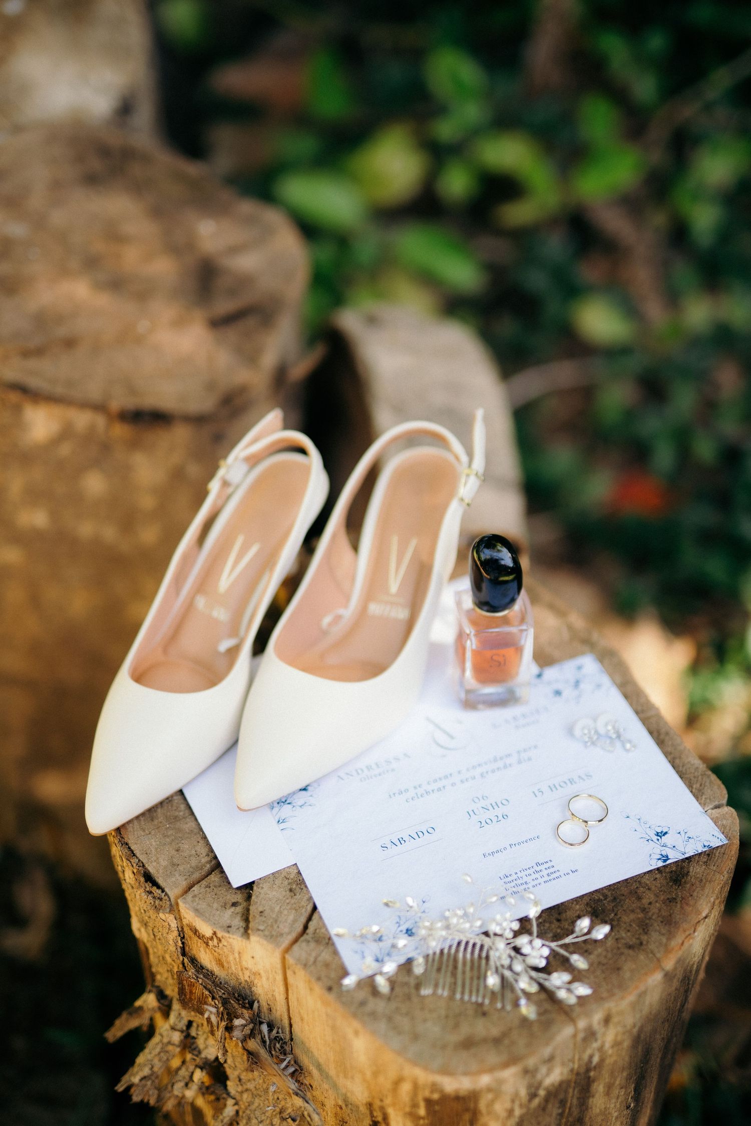 White bridal heels, wedding rings, perfume, and invitation displayed on a rustic wooden stump outdoors.