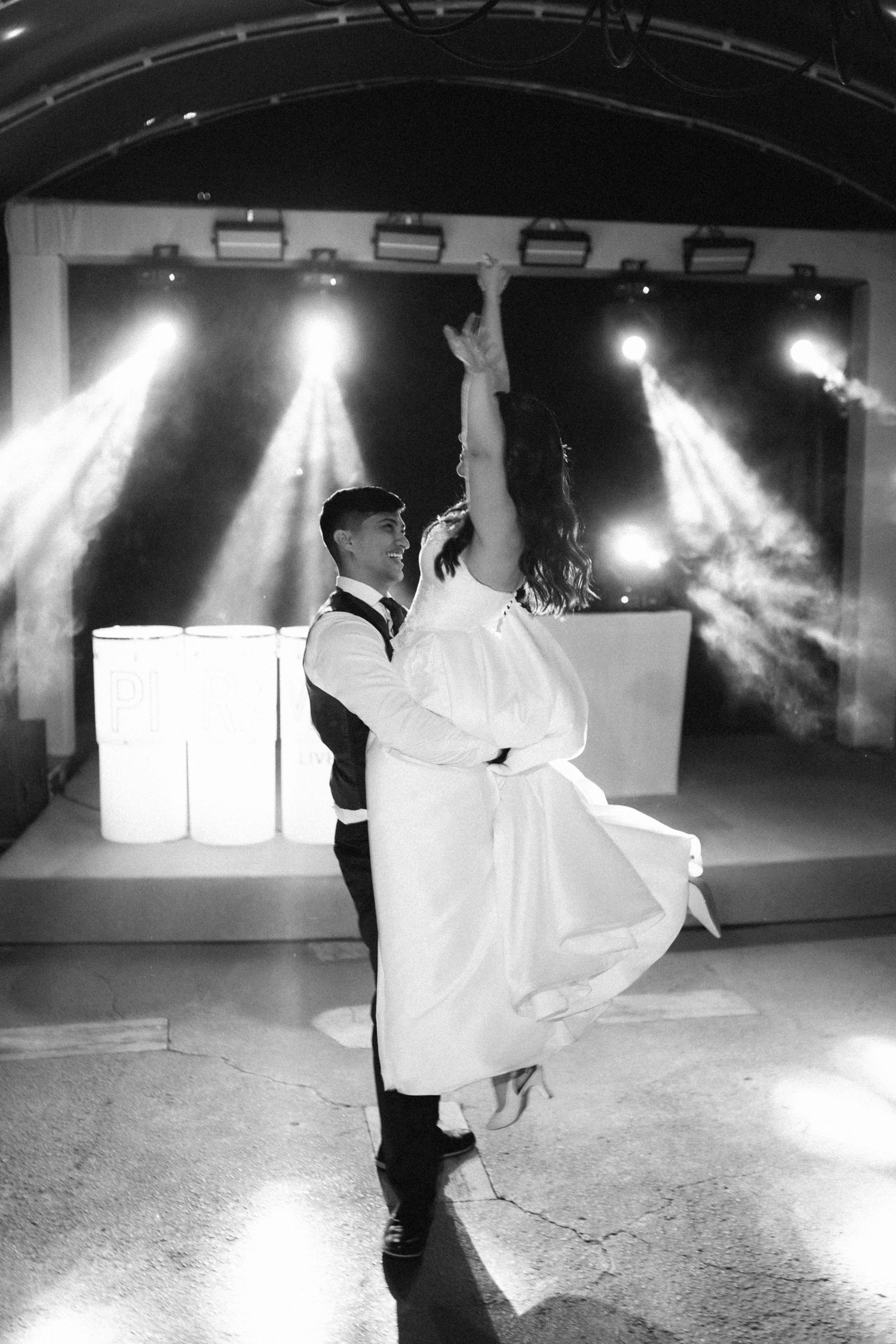 Black and white photo of a groom lifting his bride during their first dance under dramatic stage lighting.