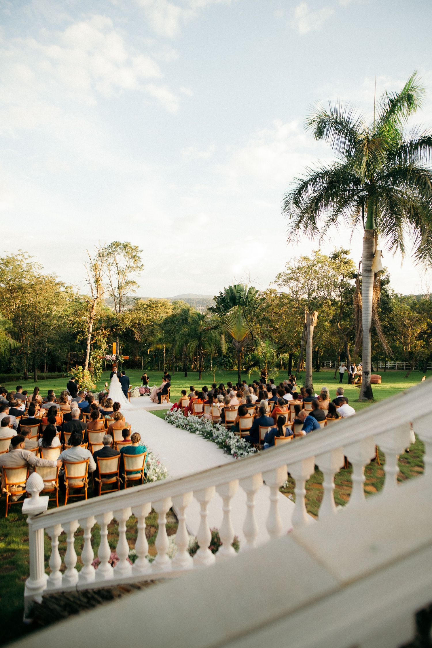 Outdoor wedding ceremony on white aisle with guests seated on wooden chairs surrounded by lush tropical palm trees.