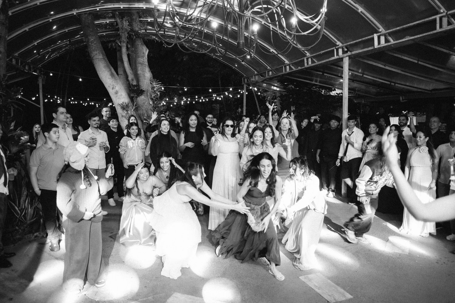 Black and white photo of bride and guests dancing at outdoor wedding reception under lights.