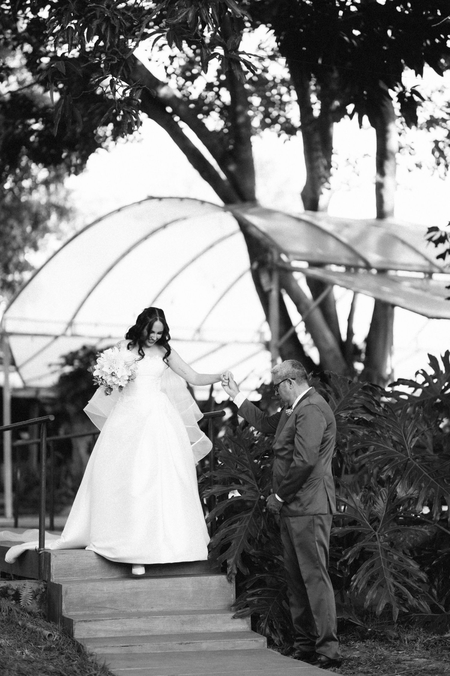 Black and white photo of a bride in a white gown descending steps with her father at an outdoor wedding.