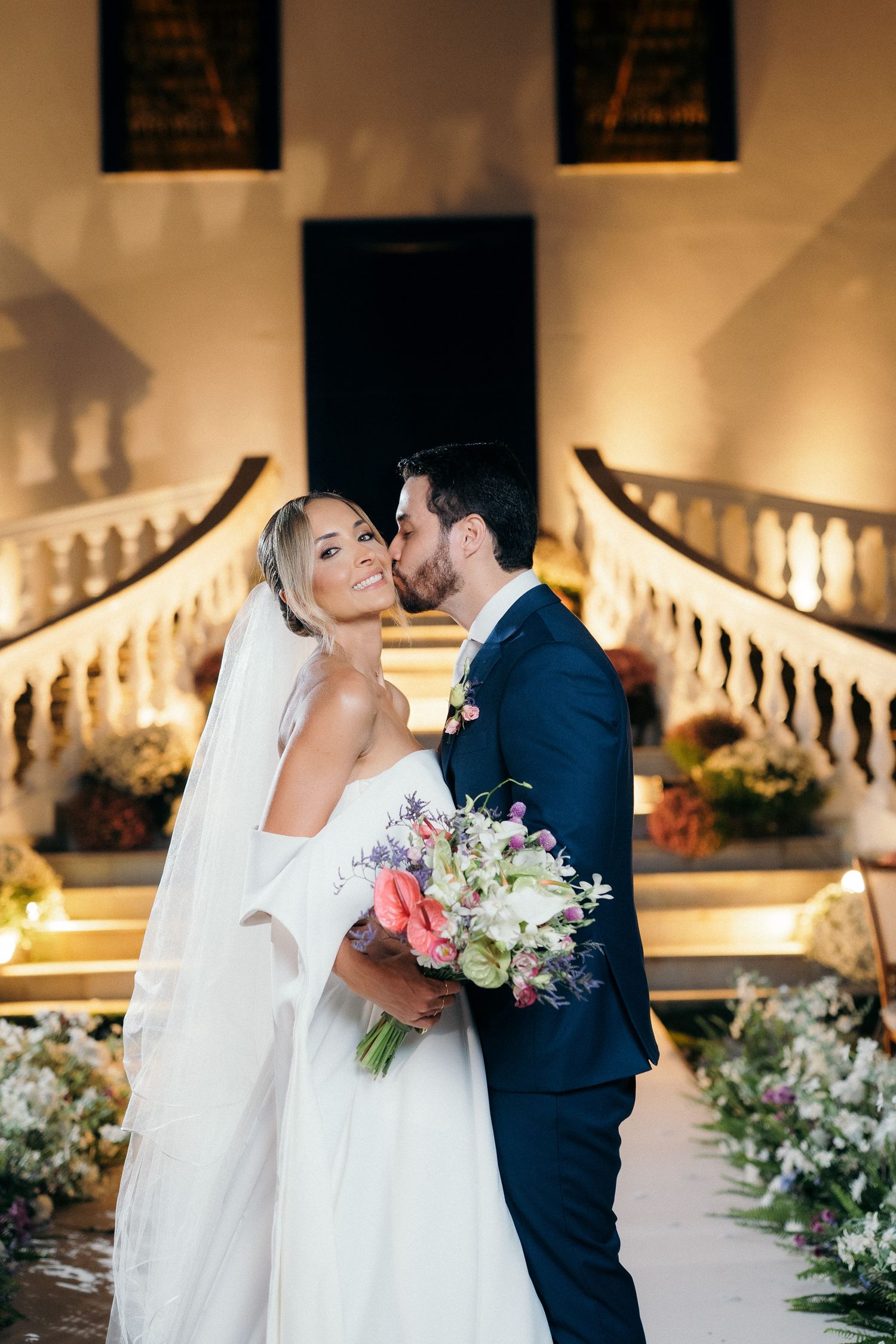 Groom kissing bride on cheek while she holds colorful bouquet on grand staircase.