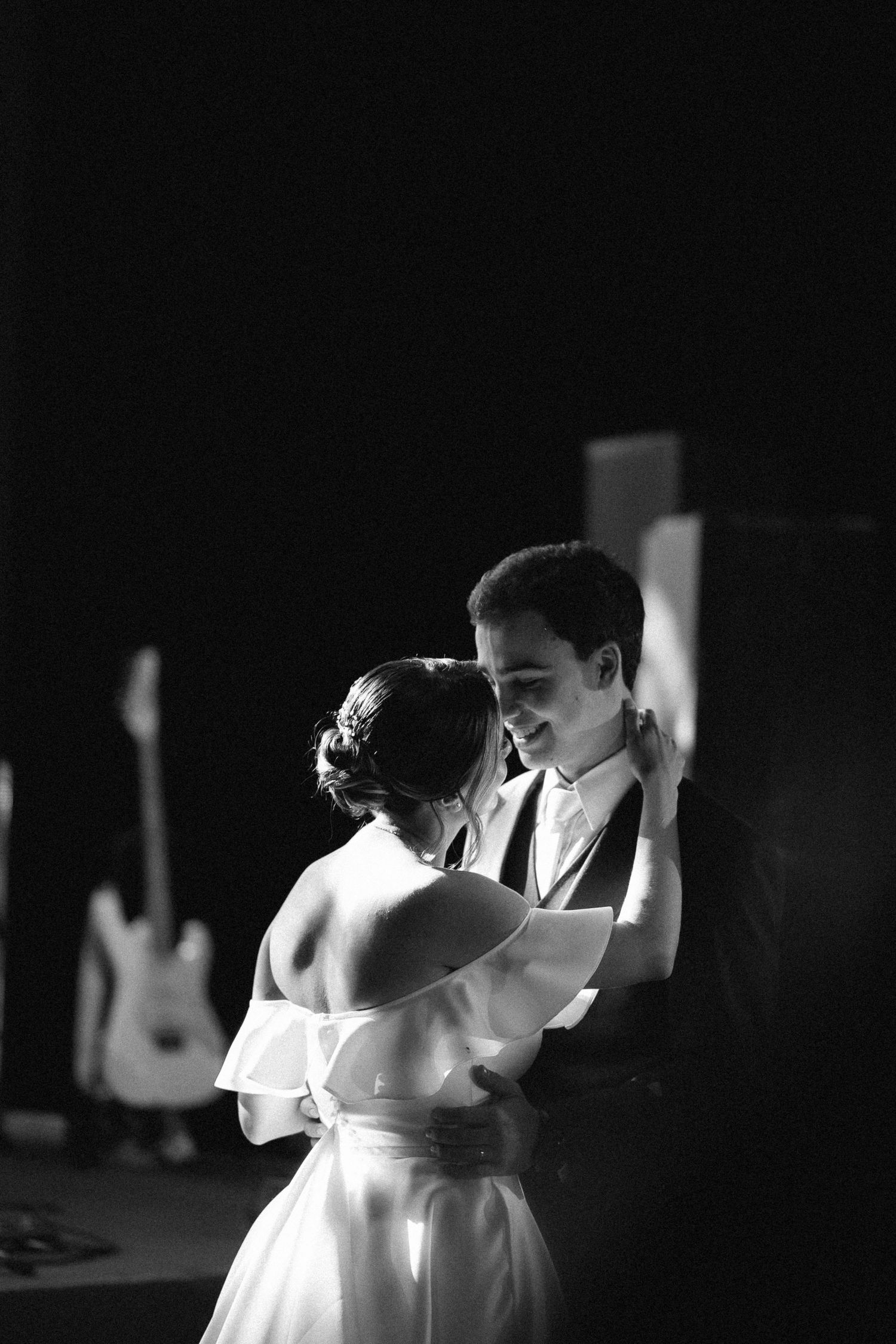 Black and white romantic photo of a bride and groom sharing their first dance with a guitar visible in background.