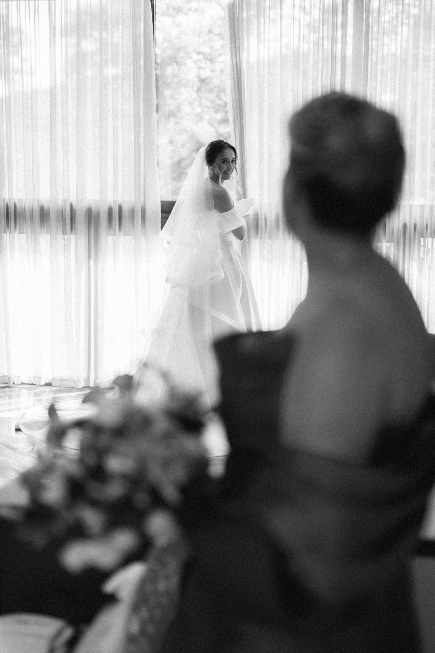 Black and white photo of a bride in a wedding gown and veil standing by sheer curtains while a guest watches.