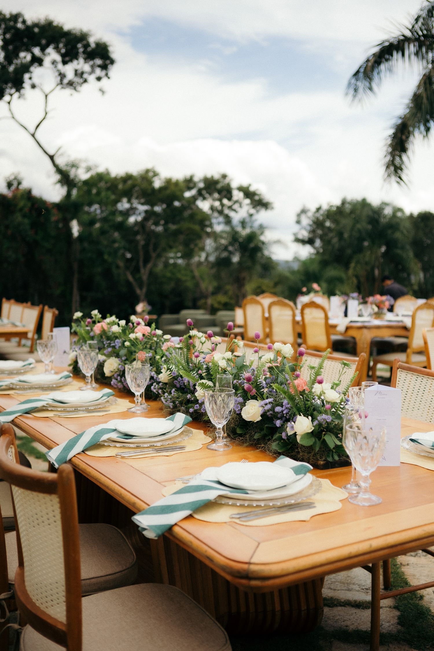 Elegant outdoor wedding reception table with floral centerpieces, crystal glasses, and teal striped napkins amid lush greenery.