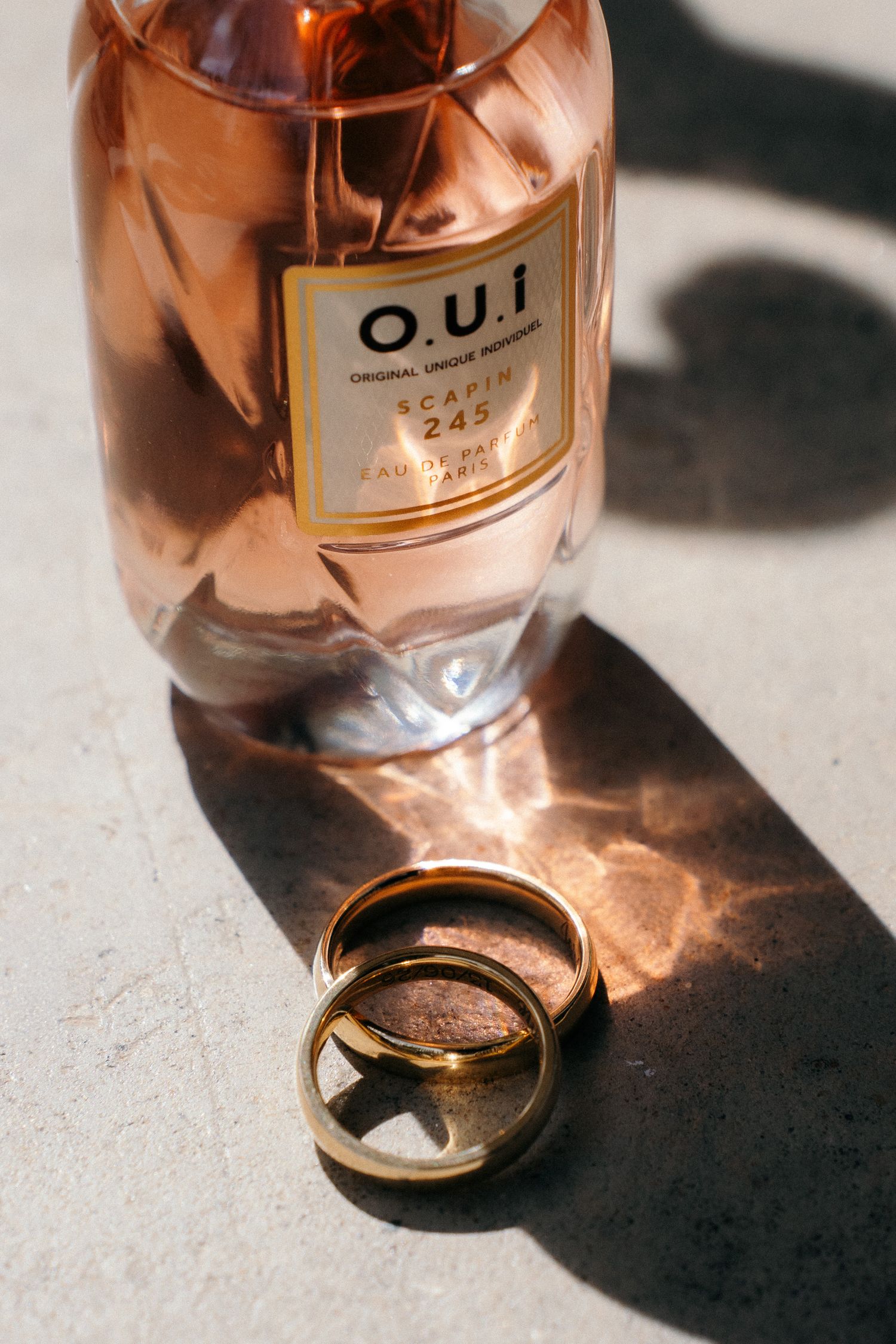 Two gold wedding bands resting beside a rose gold O.U.I perfume bottle on a concrete surface in sunlight.