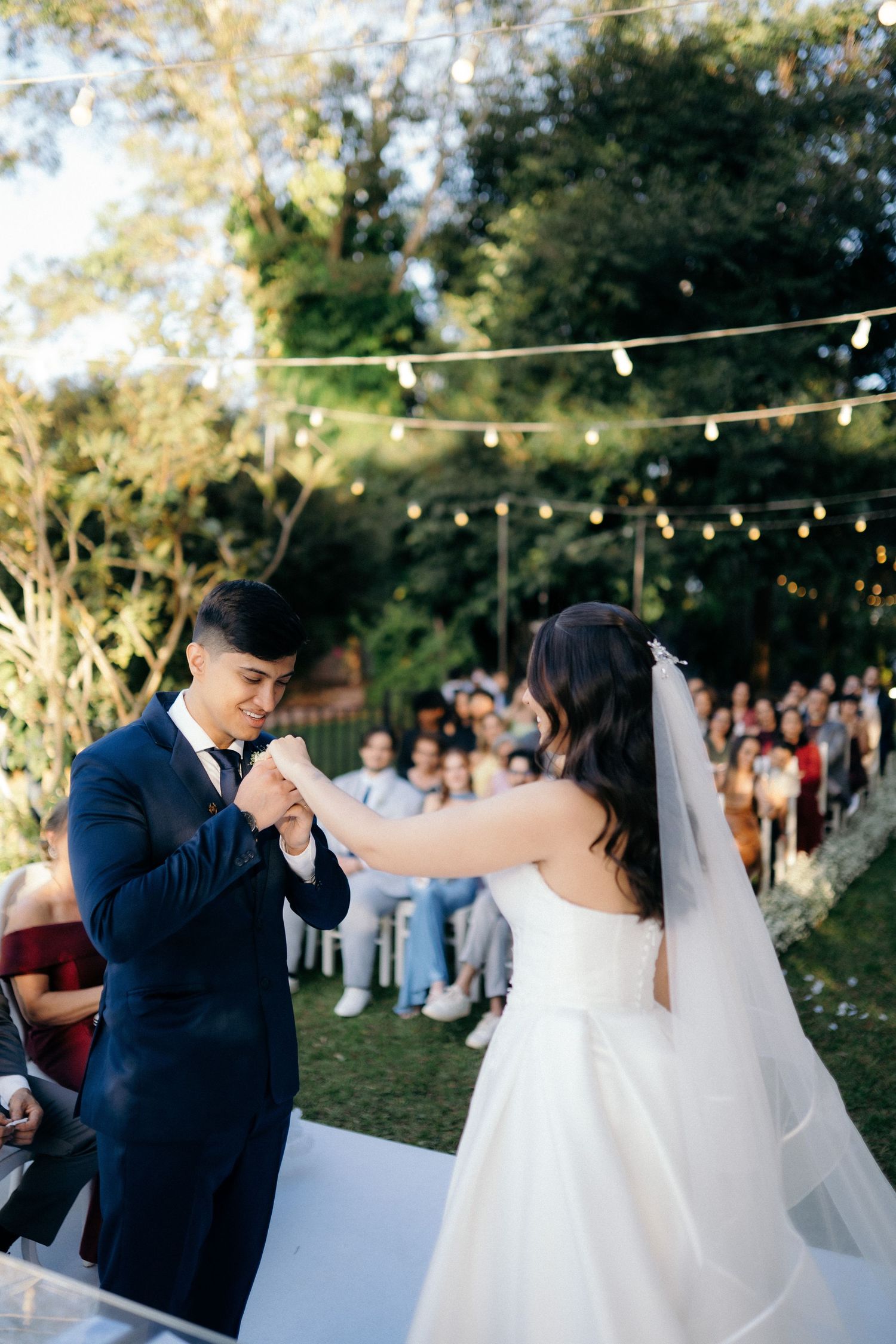 Groom in navy suit holds bride's hand at outdoor wedding ceremony with guests seated behind.