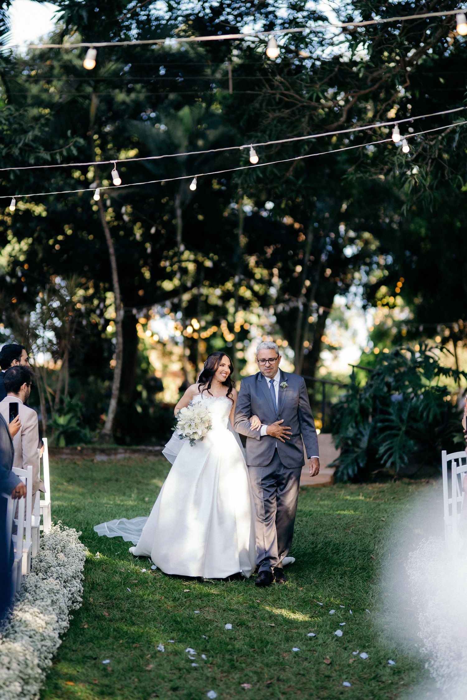 Bride walking down outdoor aisle with escort under string lights at garden wedding ceremony.