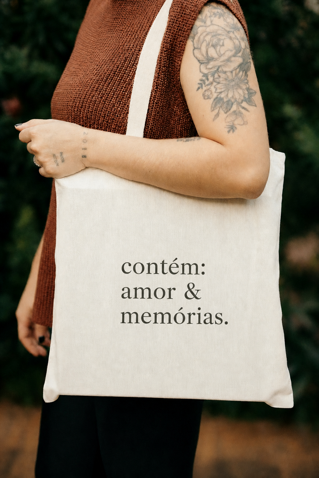 White tote bag with Portuguese text 'contém: amor & memórias' held by tattooed woman in brown knit vest.
