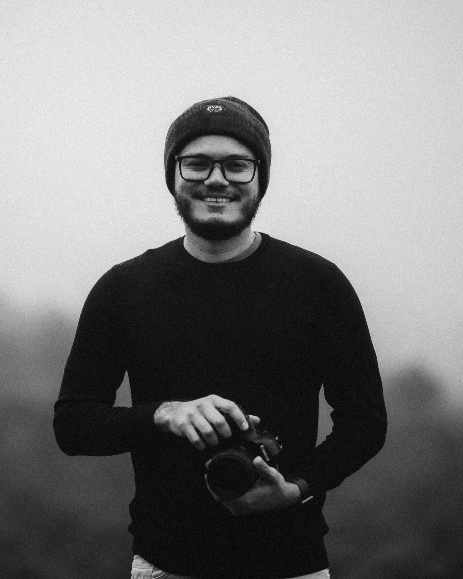 Smiling young photographer in black sweater and beanie holds camera in black and white outdoor portrait.