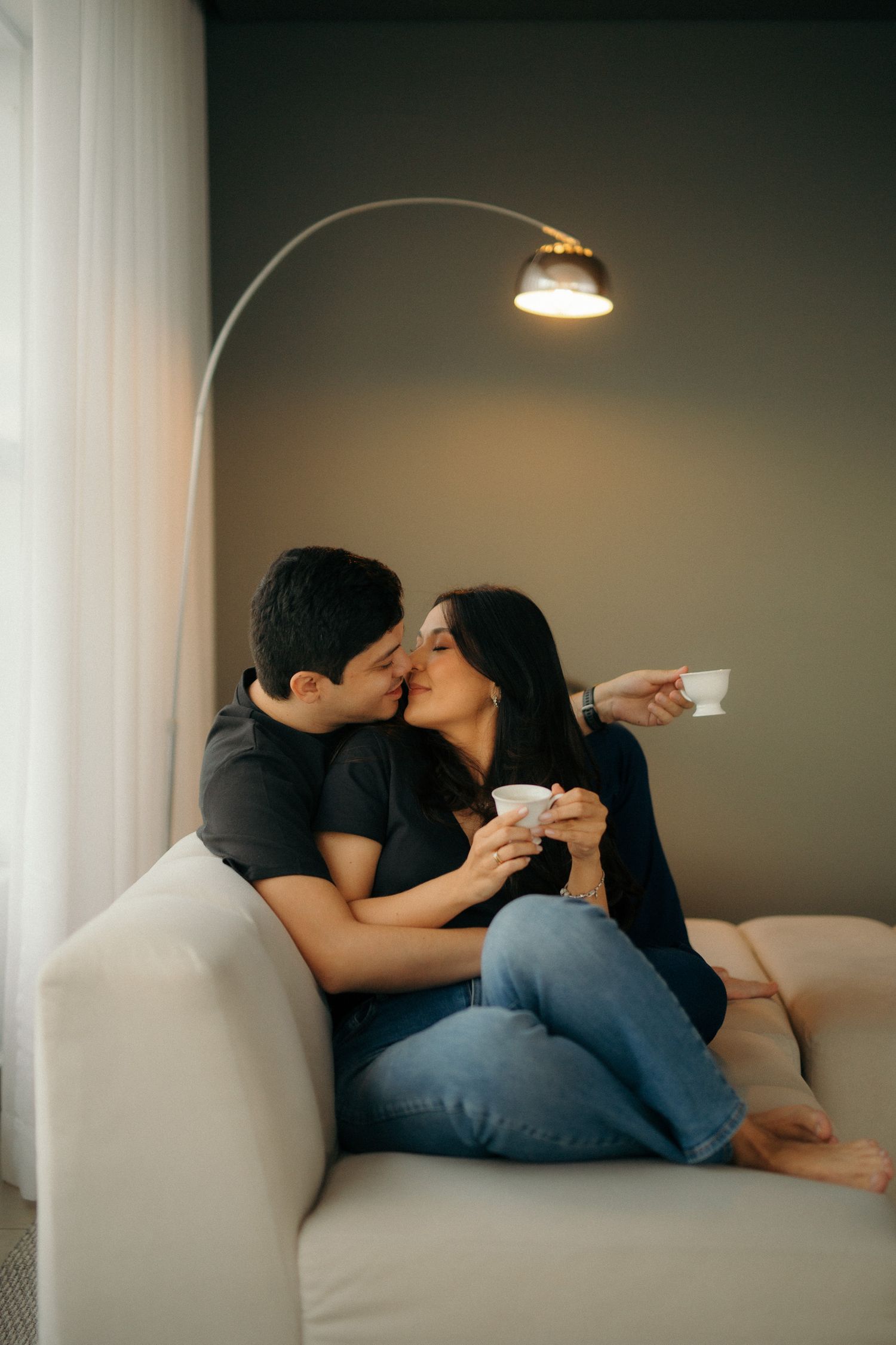 Couple kissing on a white sofa while holding coffee cups under a lamp.